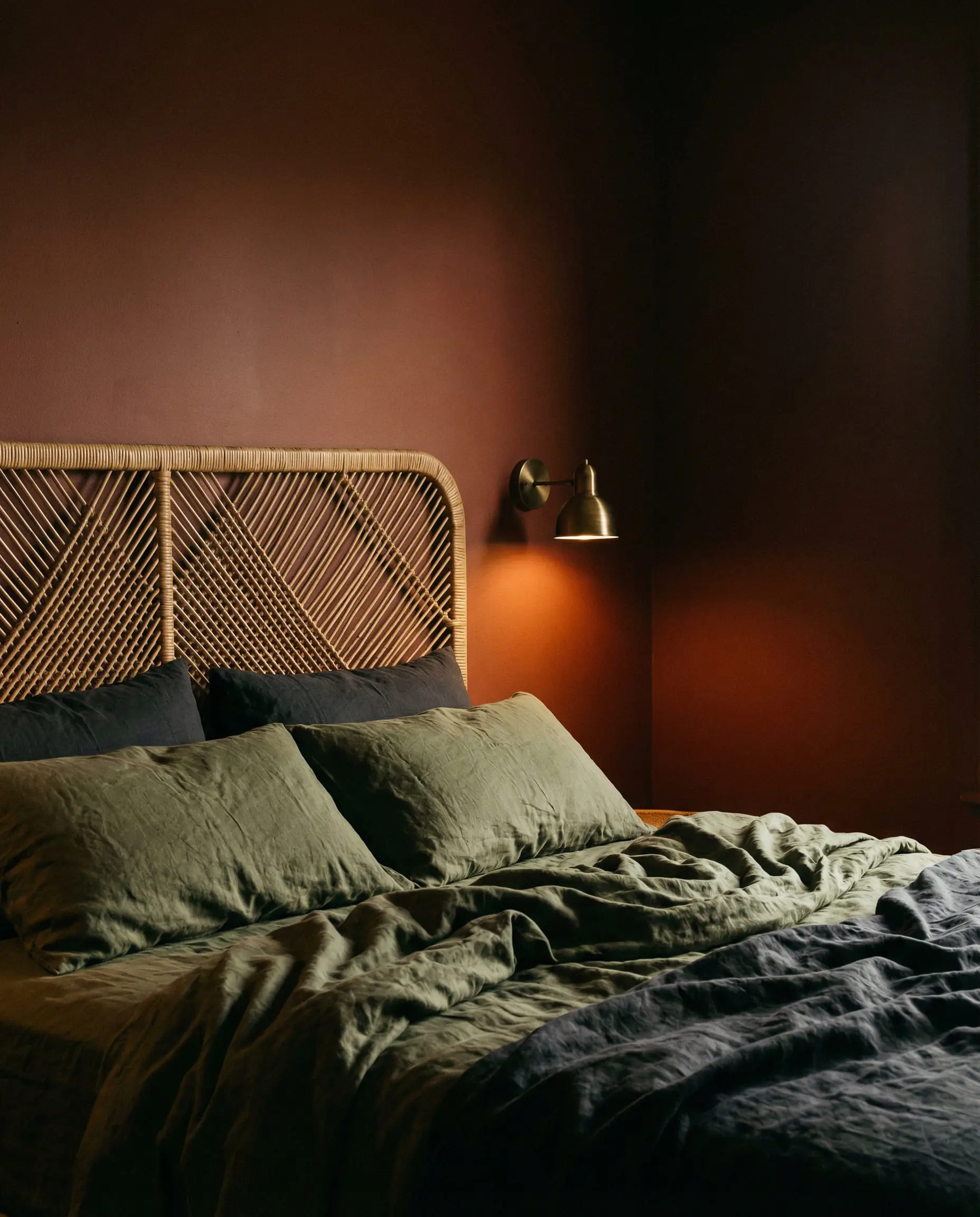 Cozy bedroom corner featuring deep rust-colored walls, a natural rattan headboard, and moody olive green linen bedding.