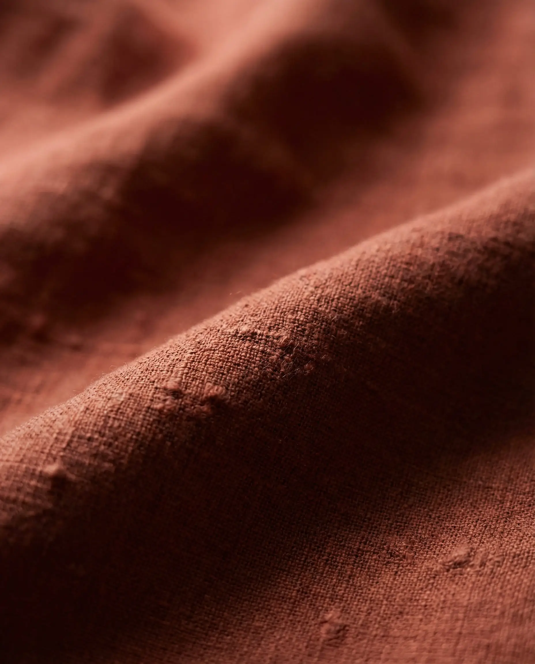 Macro detail shot of heavy washed linen fabric in a deep rust orange color showing natural weave imperfections.