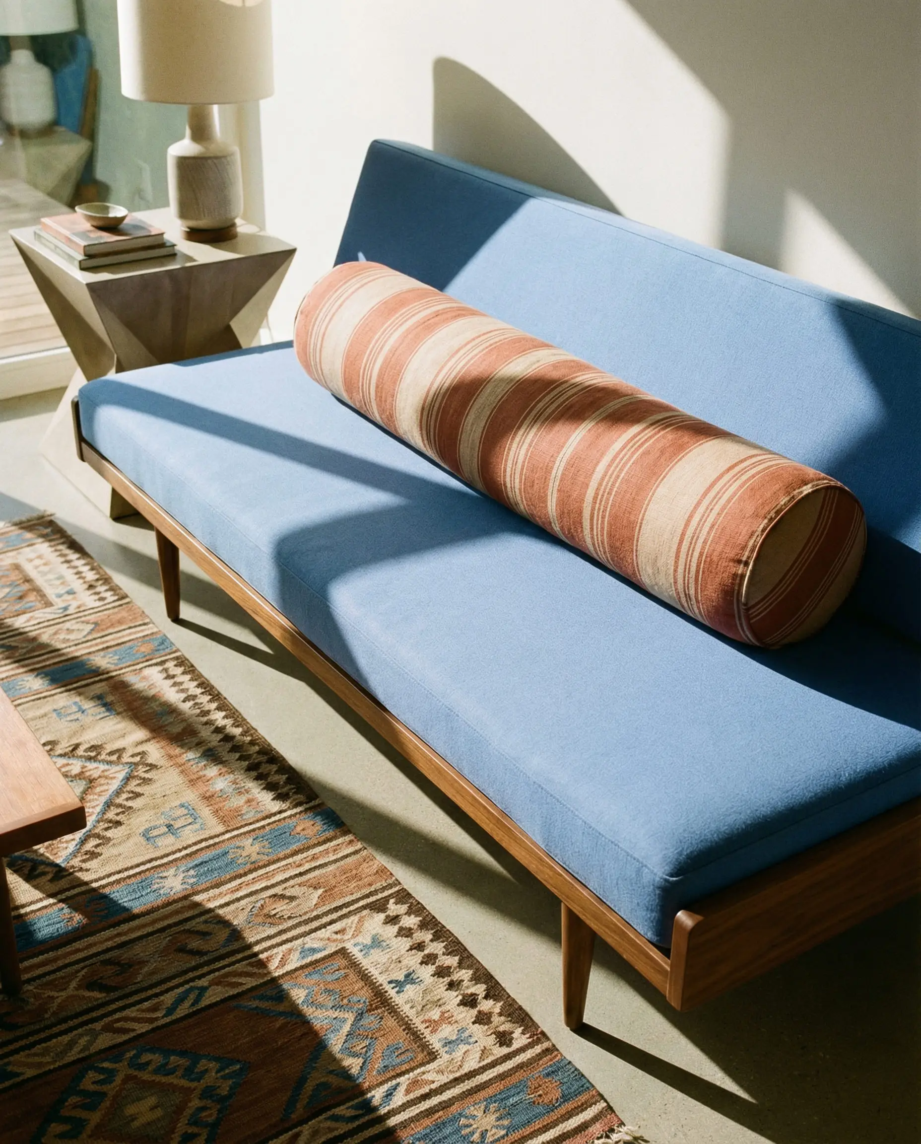A cylindrical bolster pillow in terracotta and beige stripes styling a cornflower blue daybed in a mid-century modern room.