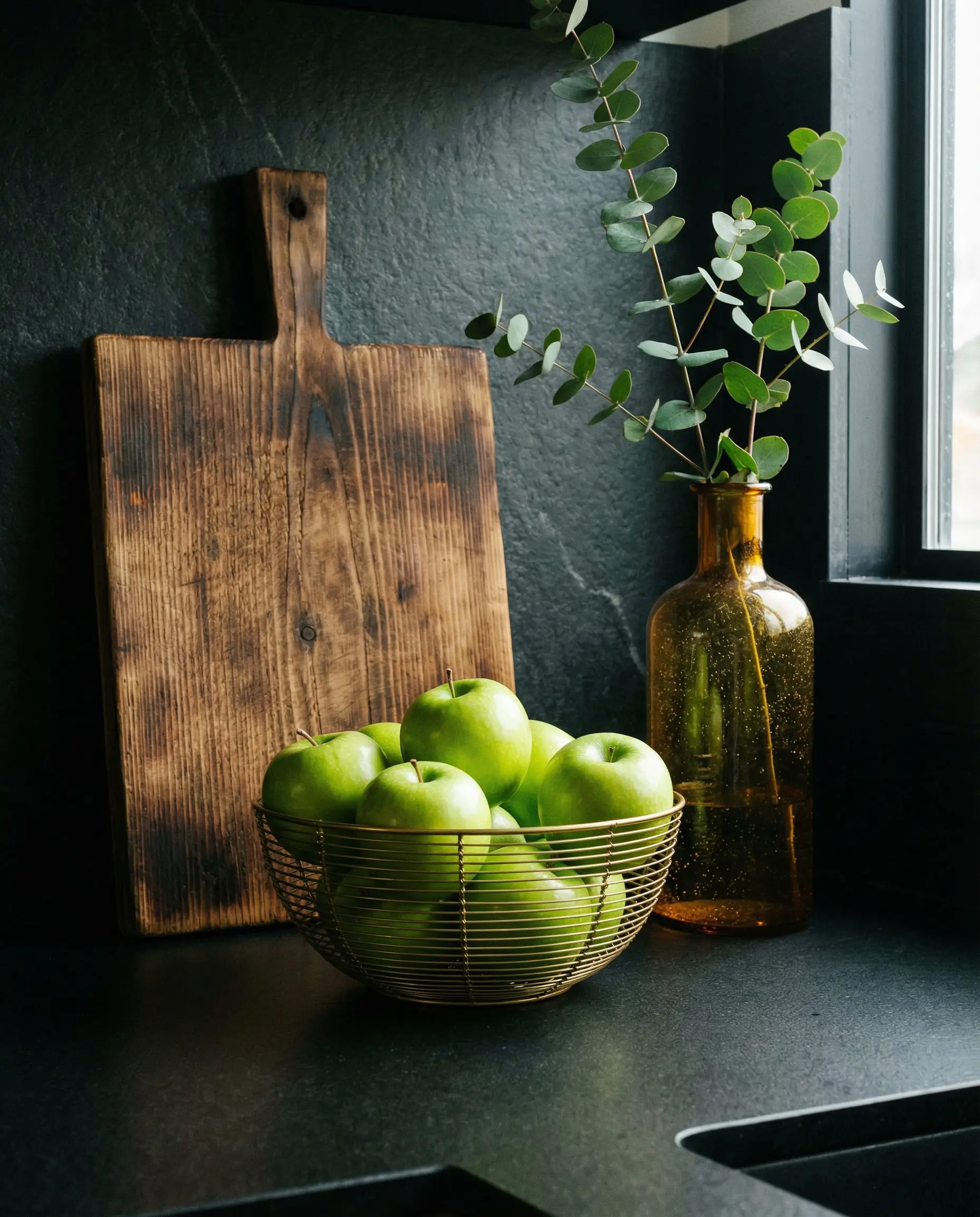 Styled kitchen vignette on black quartz with burnt wood boards, green apples in a gold bowl, and amber glass.