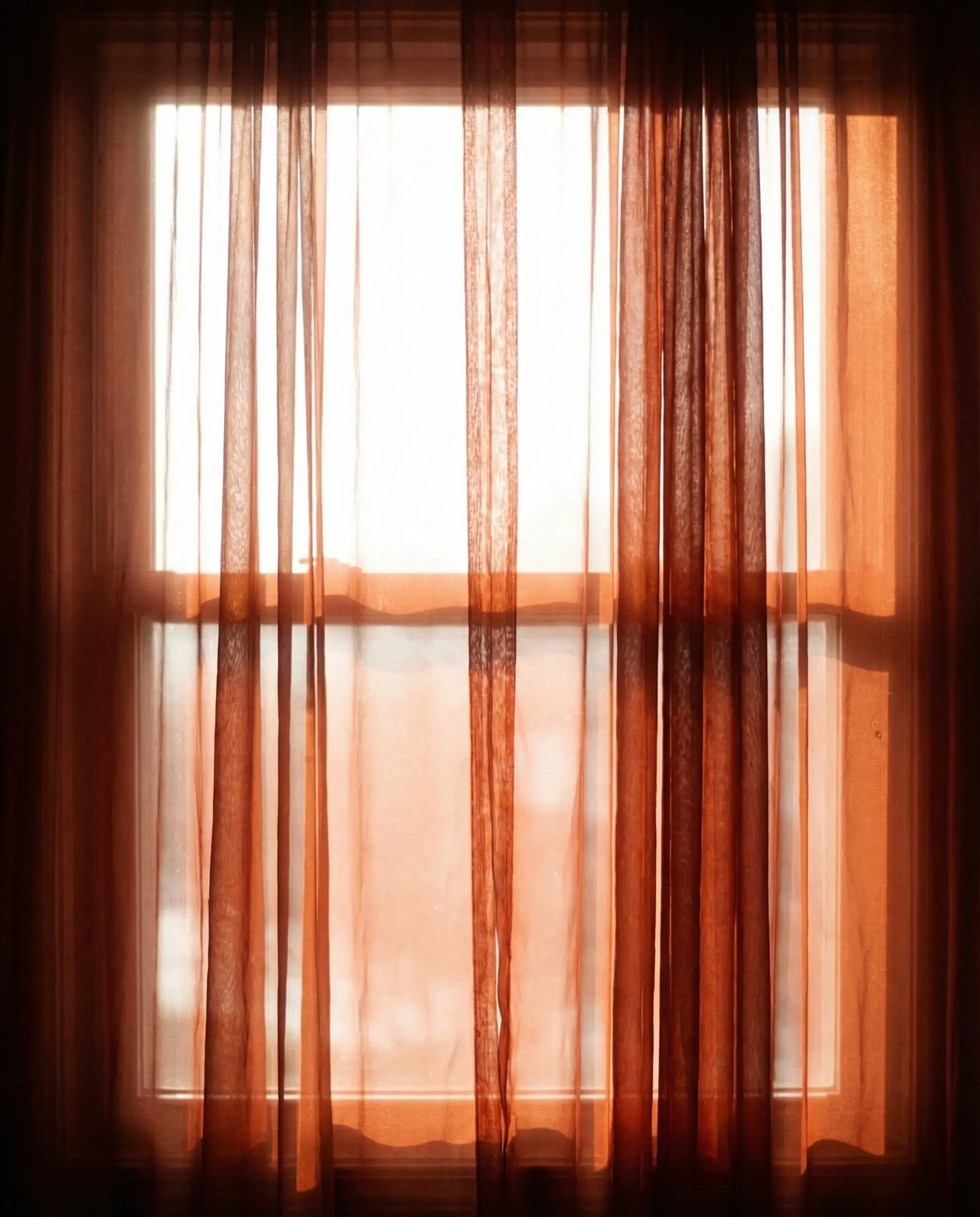 Sunlight filtering through semi-sheer rust orange voile curtains creating a warm glowing effect.