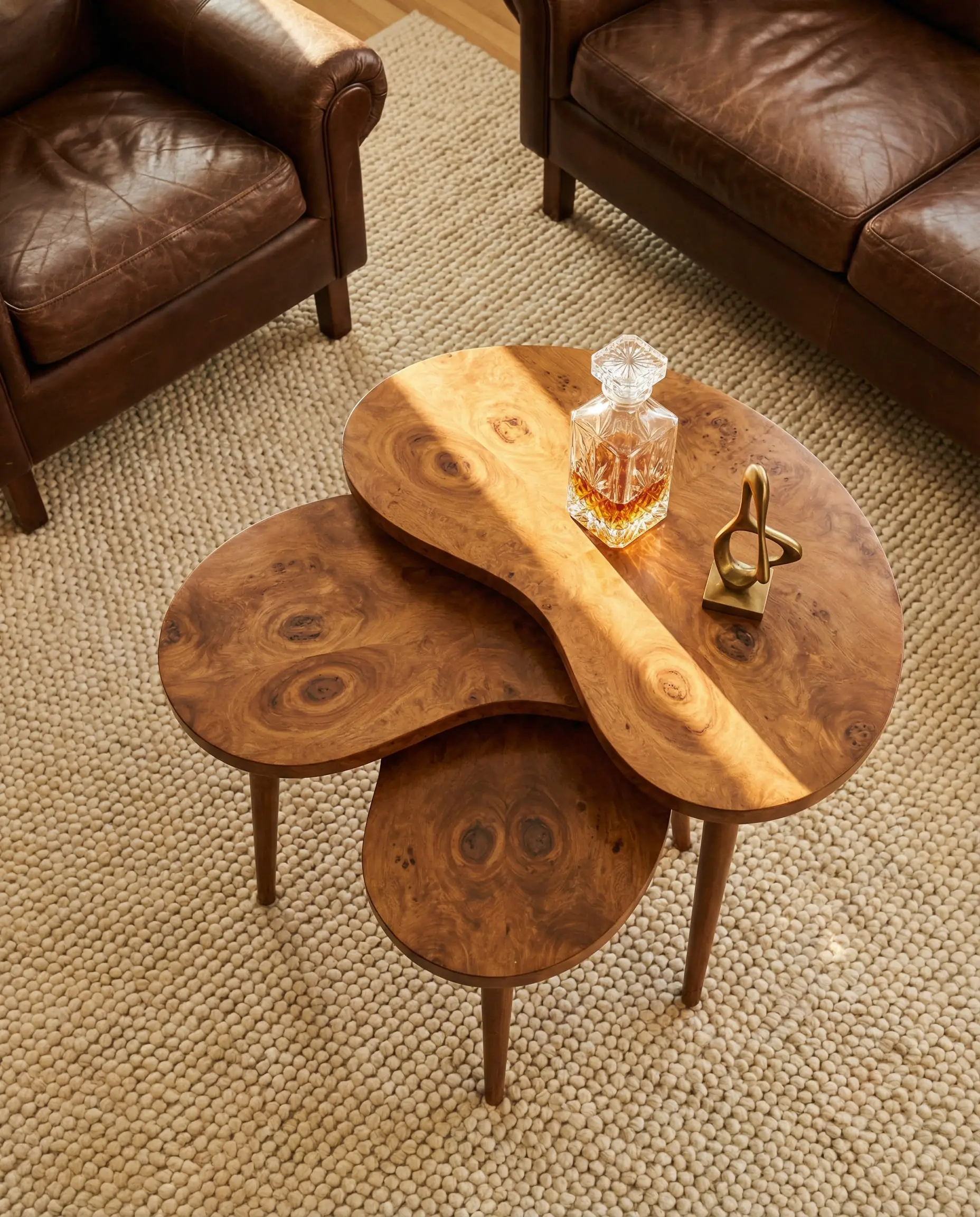 Top-down view of 1970s vintage burl wood nesting tables styled with a chocolate brown leather armchair and cream wool rug.