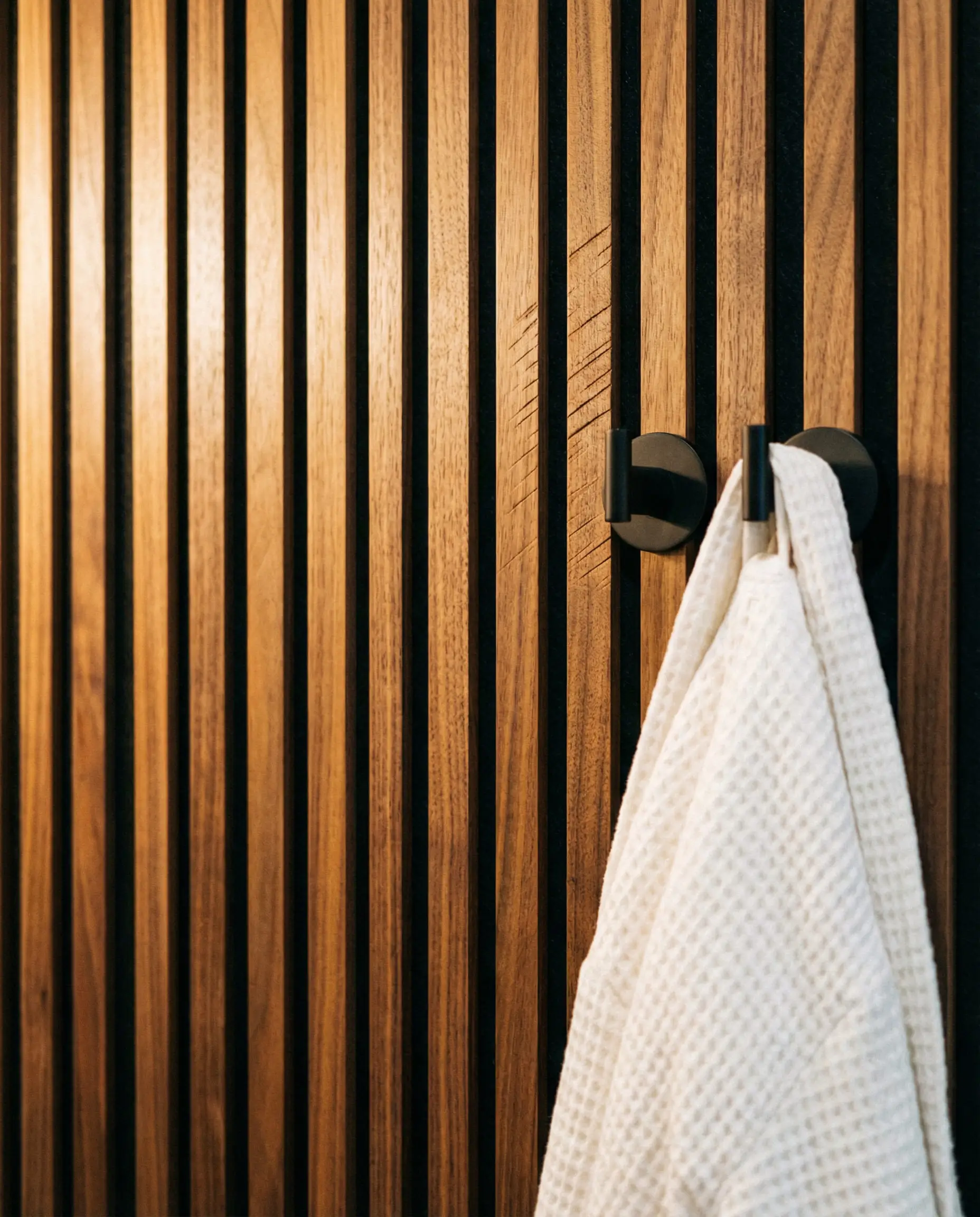 Close-up detail of vertical walnut wood slats on a bathroom wall with a black robe hook.