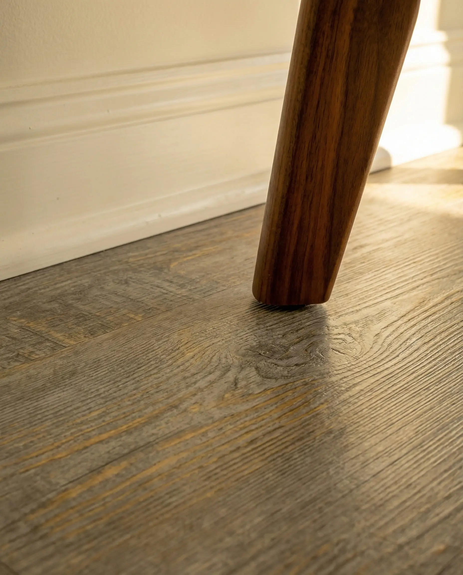 Macro close-up of warm greige luxury vinyl plank flooring with brown undertones paired with creamy off-white baseboards and walnut wood.
