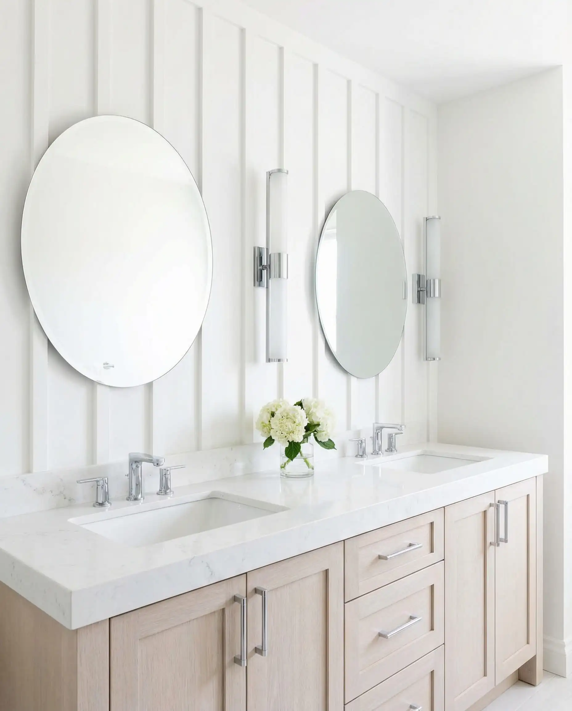 Bright bathroom vanity wall featuring white vertical board and batten with mounted round mirrors and sconces.