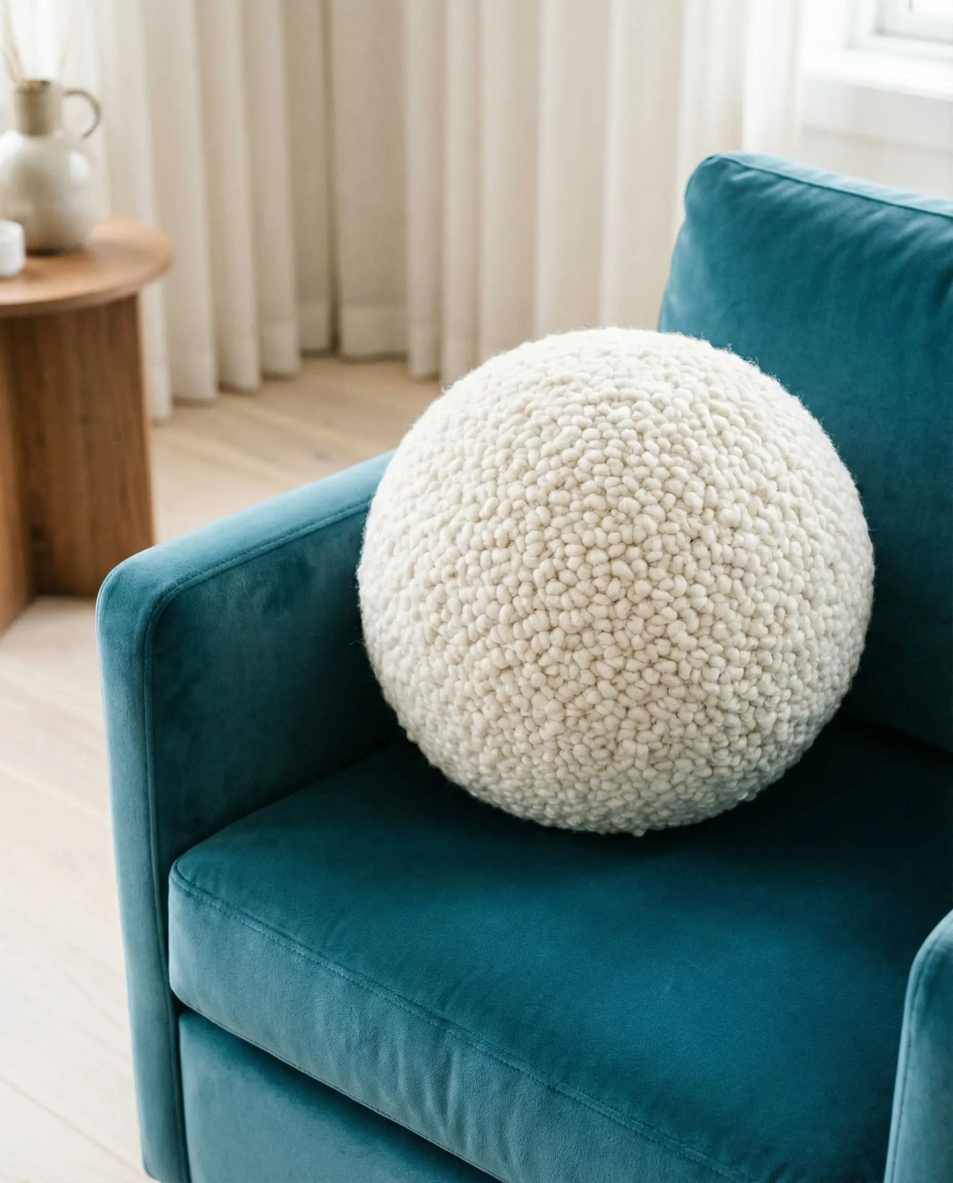 A sculptural white bouclé ball pillow resting on a teal blue velvet armchair, showcasing the 2026 spherical pillow trend.