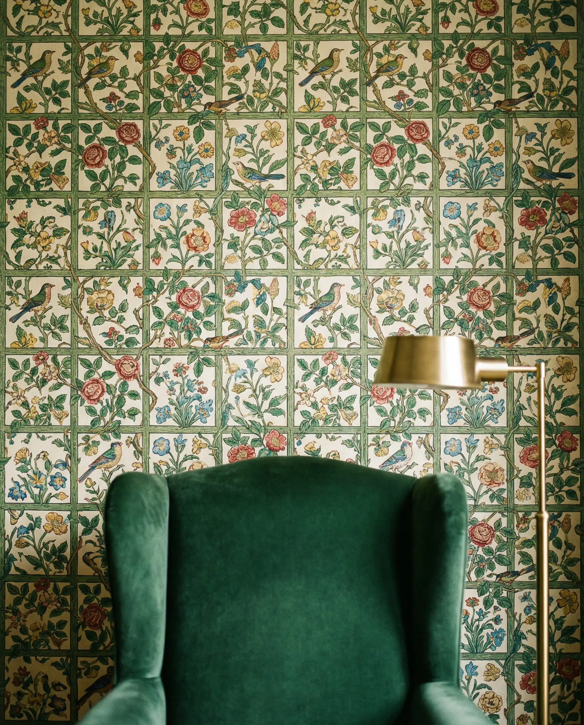 Interior wall covered in William Morris Trellis vine wallpaper with a green velvet chair in the foreground.