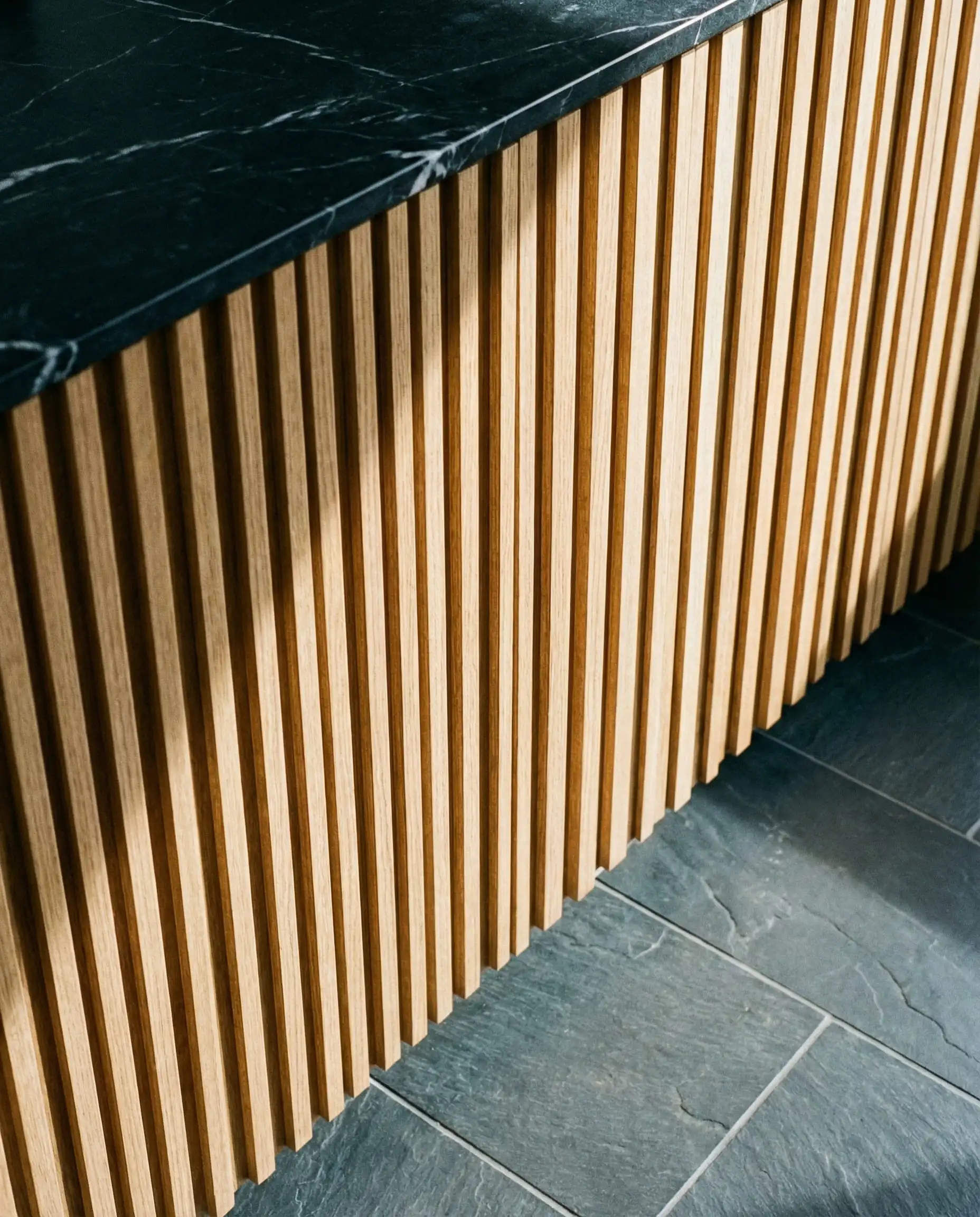 Vertical white oak wood slat cabinetry detail against a dark gray slate tile floor.
