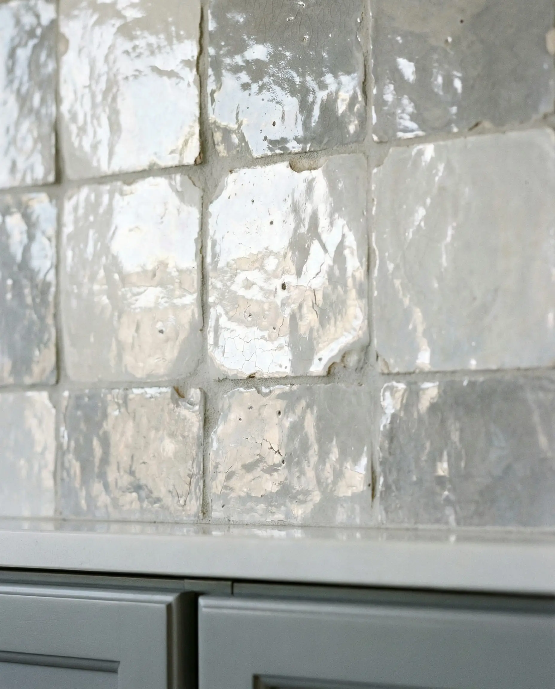 Macro detail of pearlescent white and gray Zellige tiles showing glossy, imperfect texture above a gray cabinet.