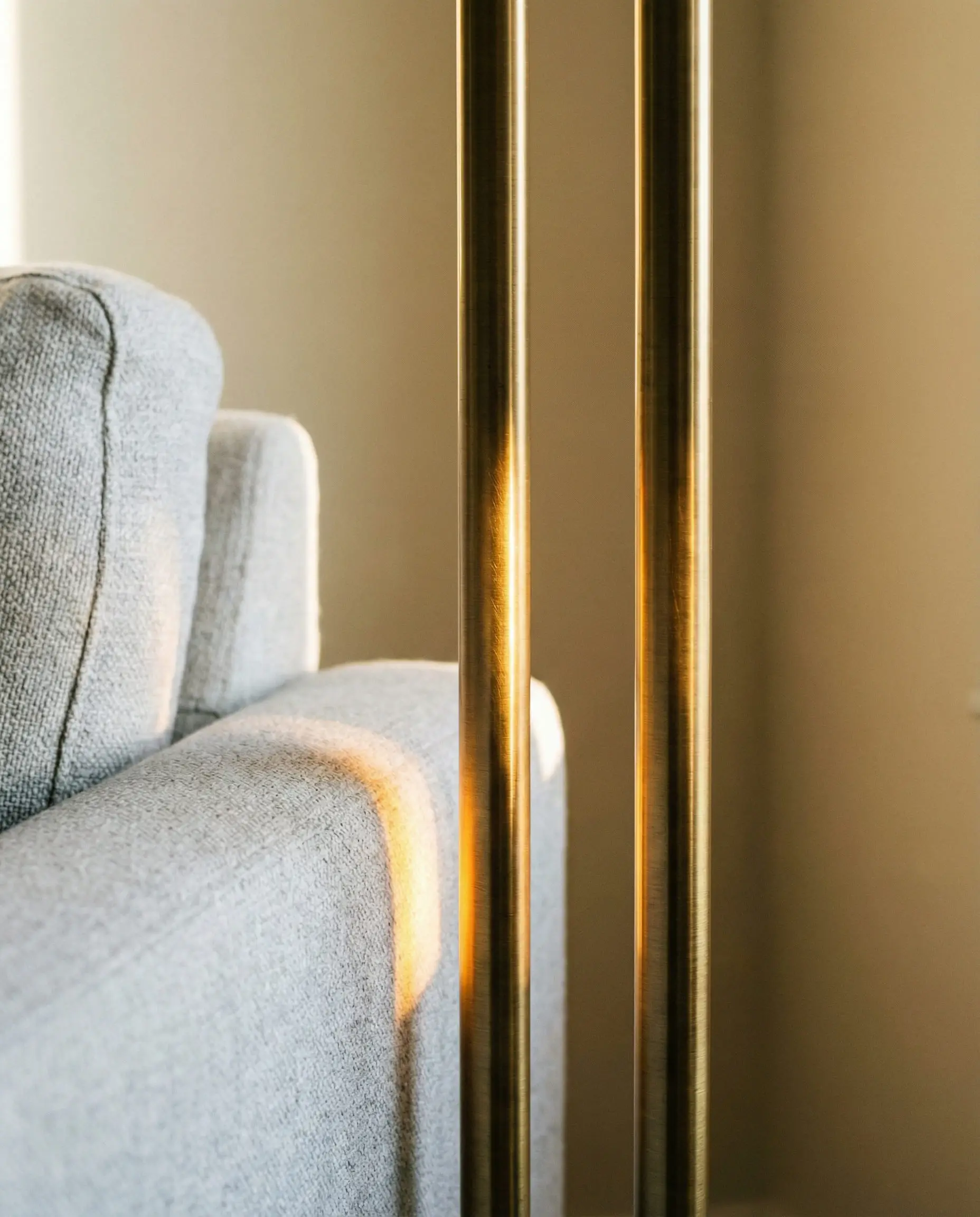 Interior styling close-up of an antique brass floor lamp stand next to a light grey sofa arm against a beige wall.