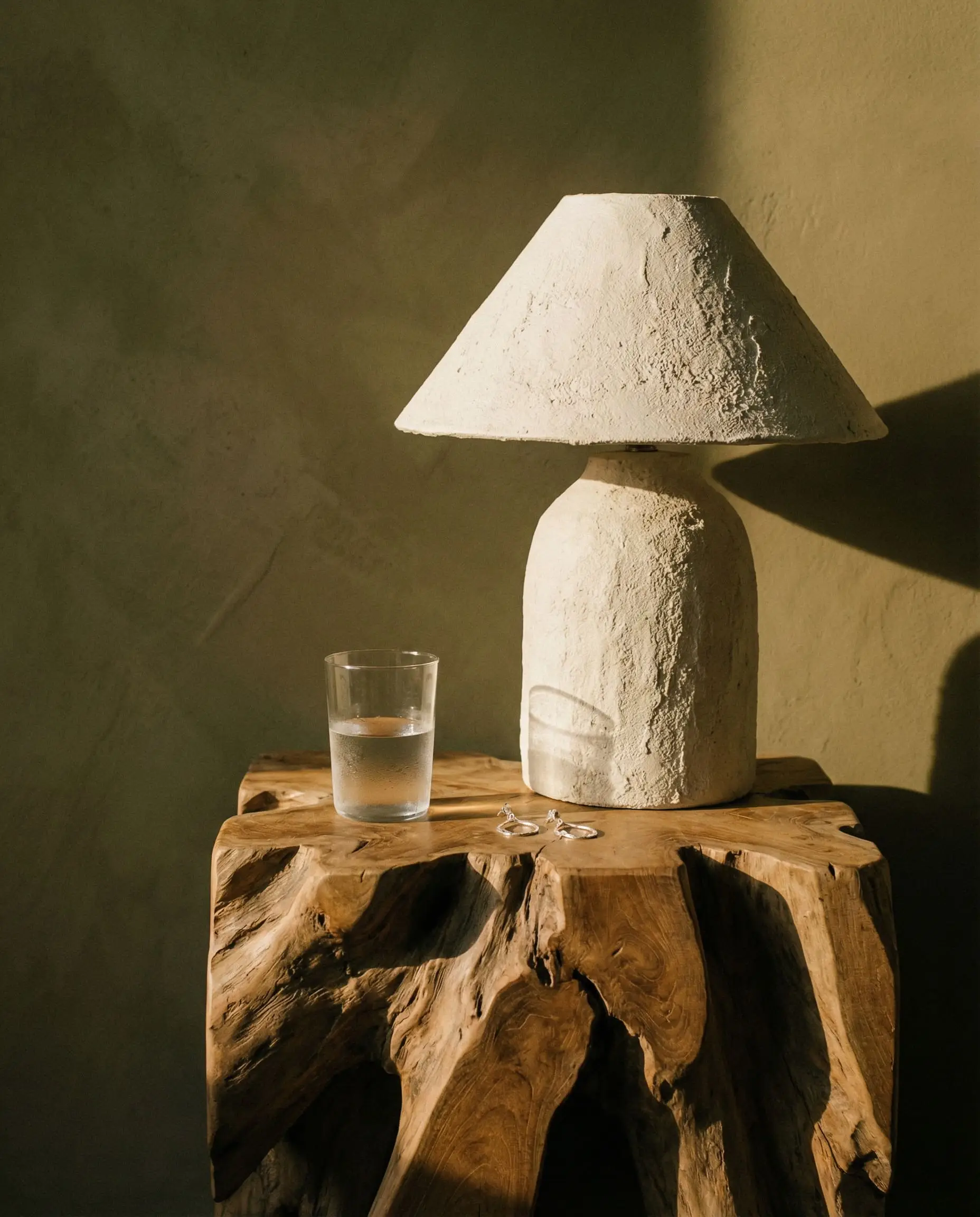 An asymmetrical raw reclaimed wood nightstand holds a sculptural wabi-sabi textured plaster table lamp against a muddy olive green limewash wall.