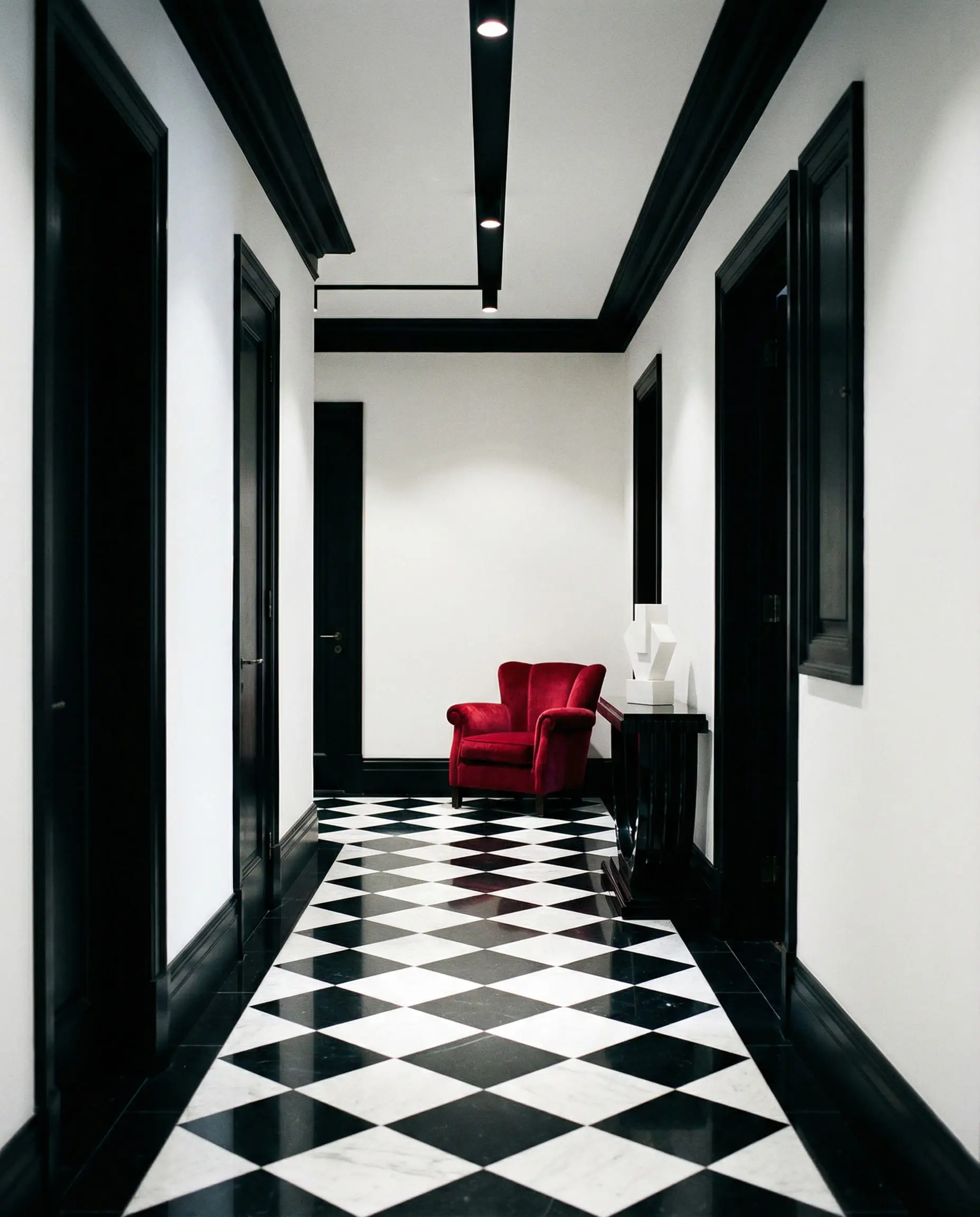 High-contrast hallway with white walls, black trim, checkerboard floors, and a single ruby red velvet accent chair.