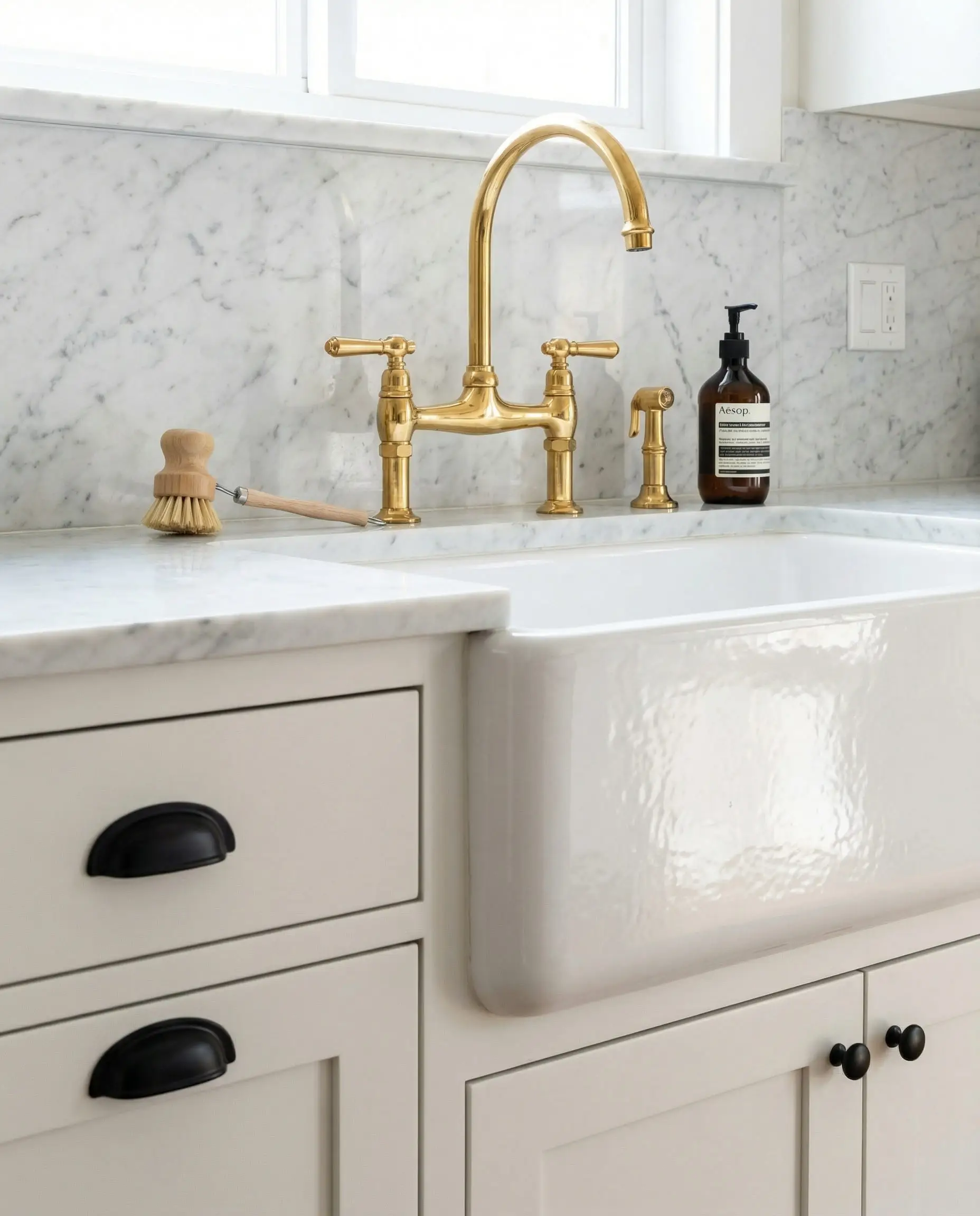 Kitchen sink area showing matte black cabinet hardware on lower doors juxtaposed with a polished unlacquered brass bridge faucet and marble backsplash.