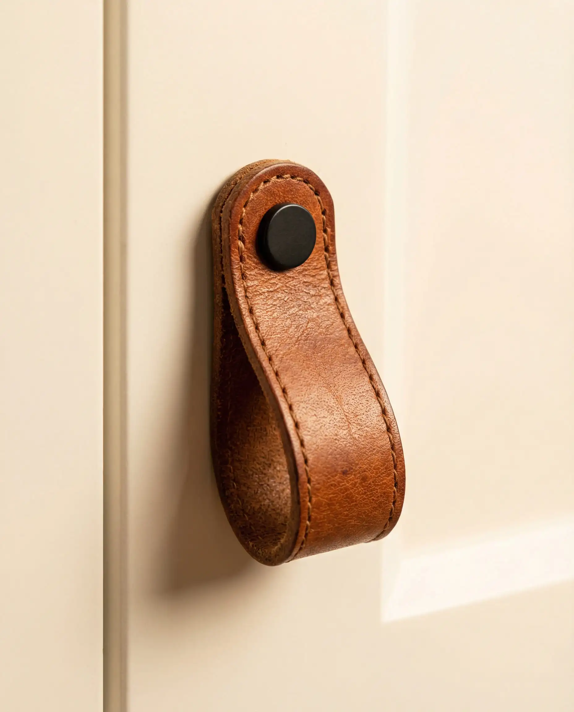 Macro shot of a black leather-wrapped cabinet handle with stitching details mounted on a warm white slab door.