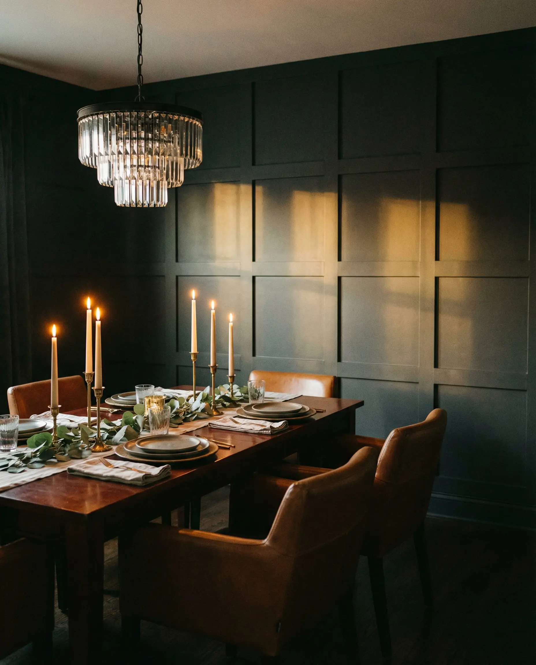 Moody dining room featuring a dark charcoal grid-patterned board and batten wall lit by candlelight and a crystal chandelier.