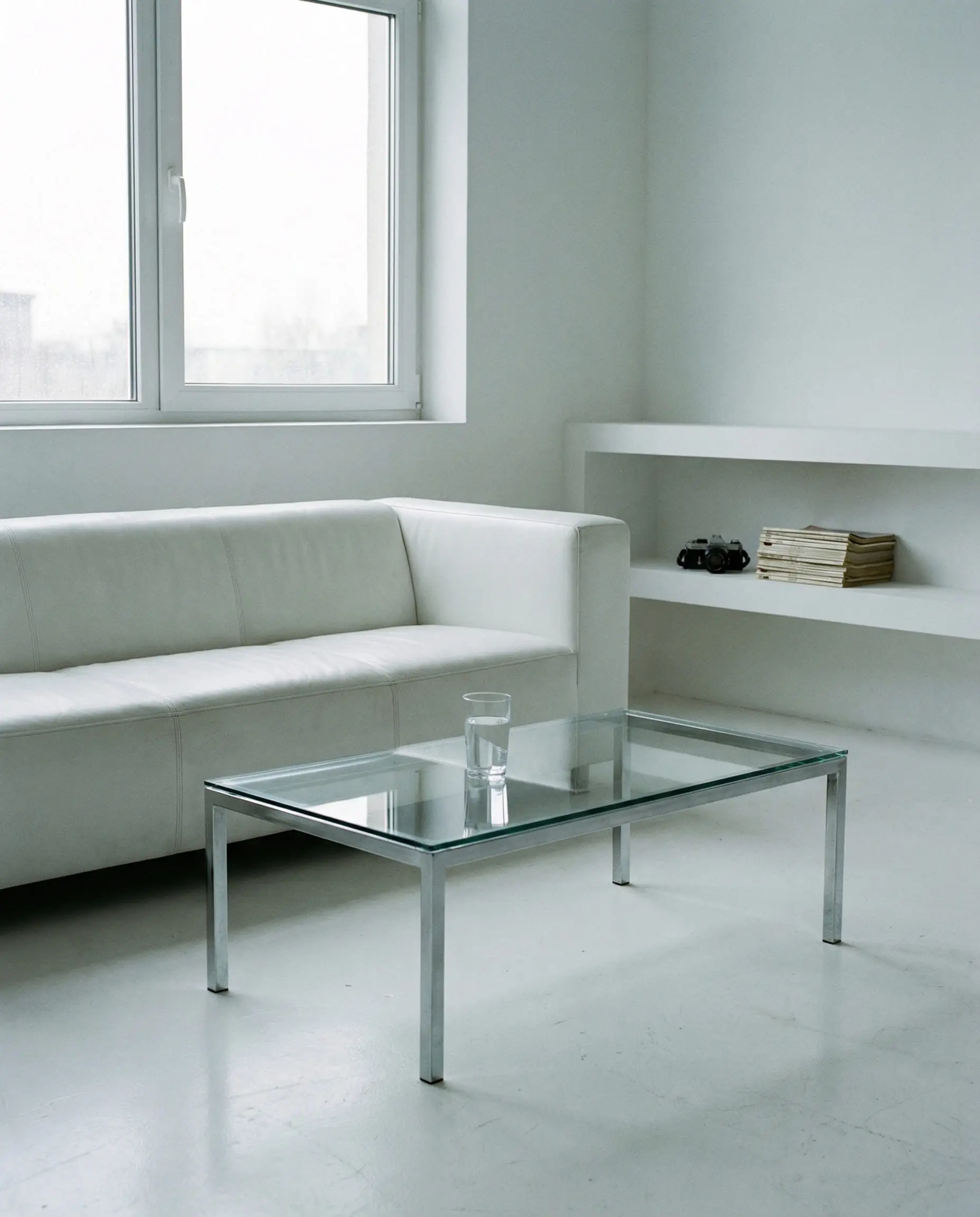 A stark and cold minimalist living room displays a boxy white leather sofa, glossy floors, and a glass coffee table lacking organic modern interior design elements.