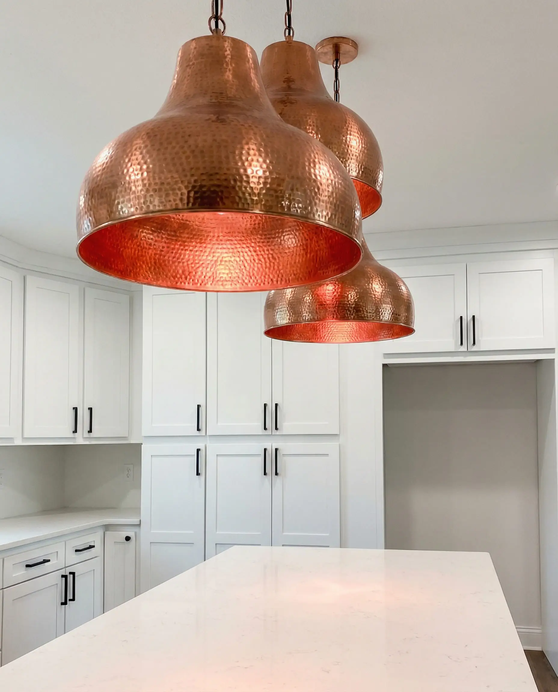 Kitchen scene showing hammered copper pendant lights hanging over an island with white cabinets and black hardware.