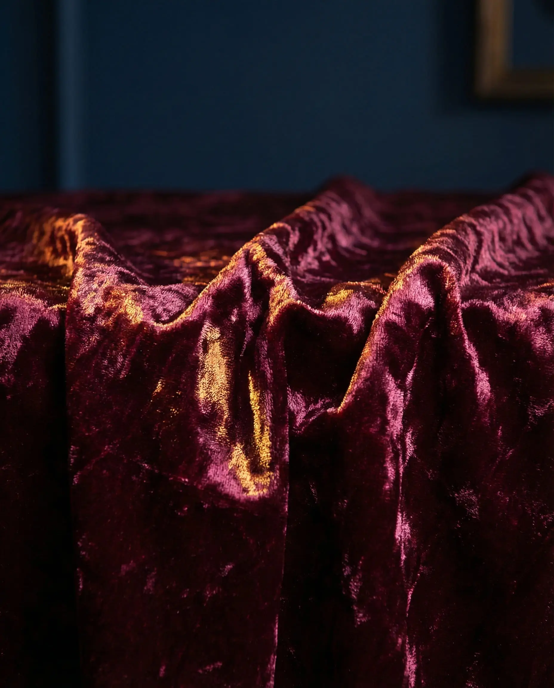 Close up of burgundy crushed velvet curtain fabric against a navy blue wall.