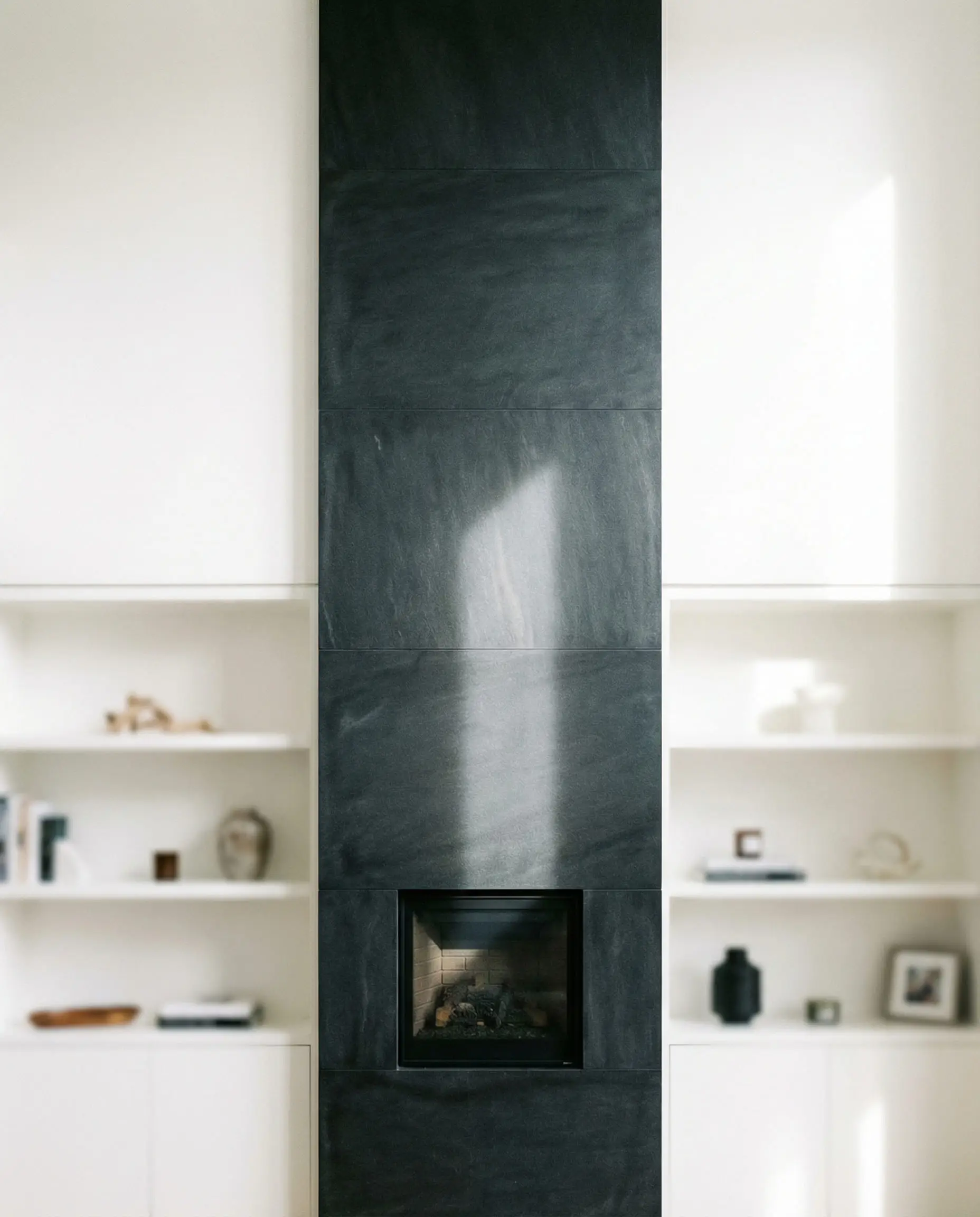 A narrow, vertical architectural column clad in dark basalt stone housing a small square fireplace insert.
