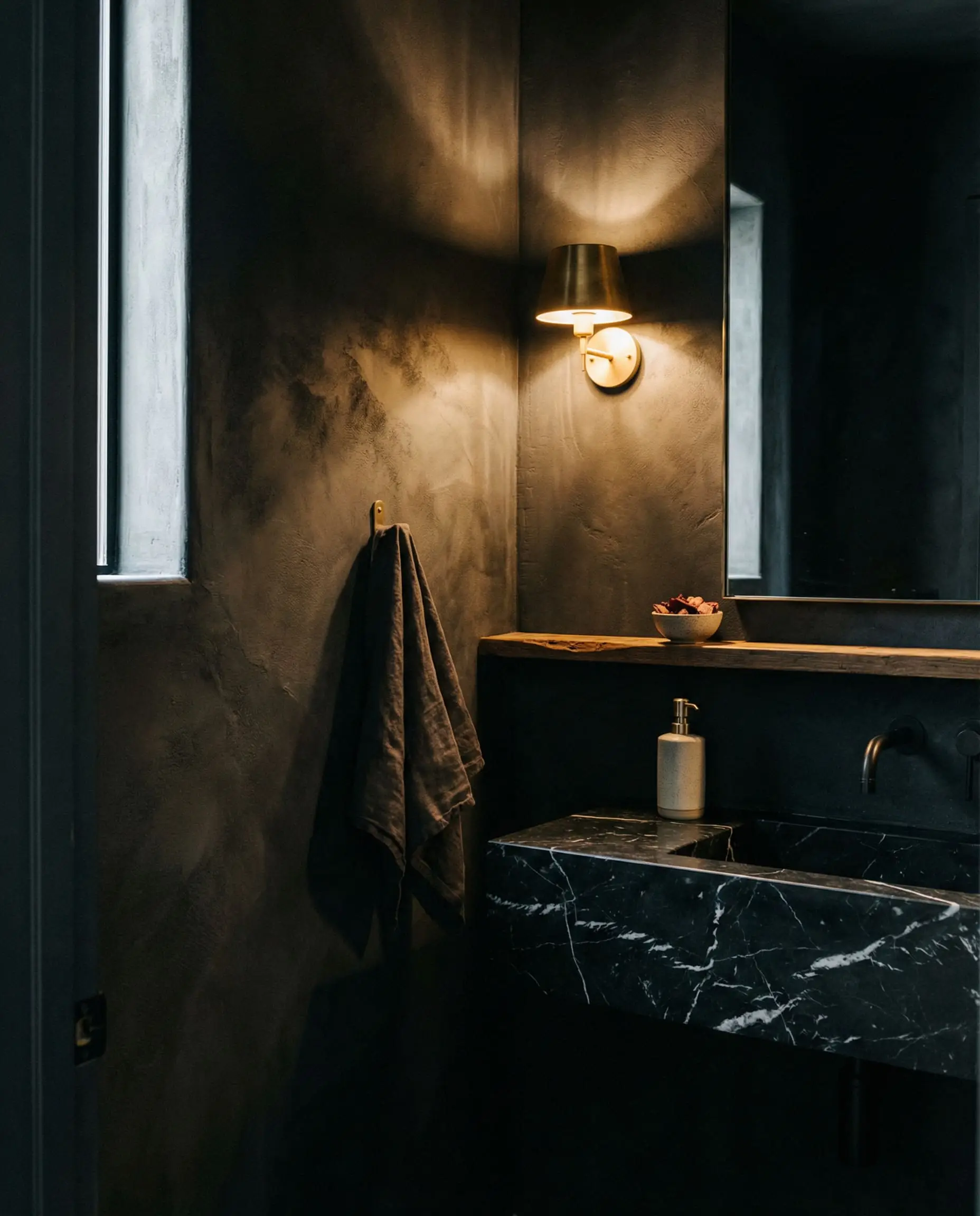 Deep charcoal roman clay colors color-drench a moody wabi-sabi powder room featuring a floating Nero Marquina marble sink and glowing brass sconce.