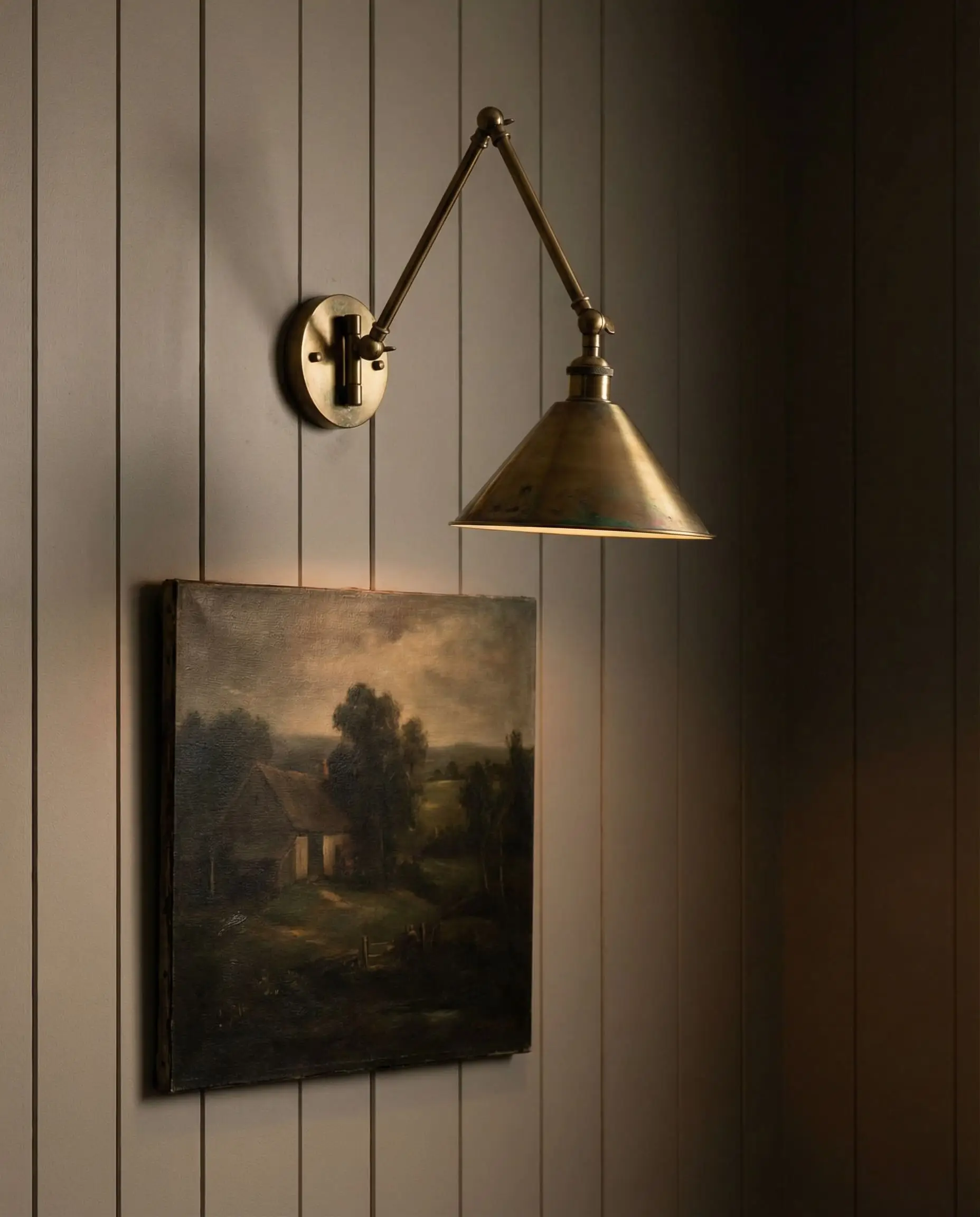 An unlacquered brass wall sconce illuminates a vintage landscape painting on a taupe European farmhouse V-groove wall.