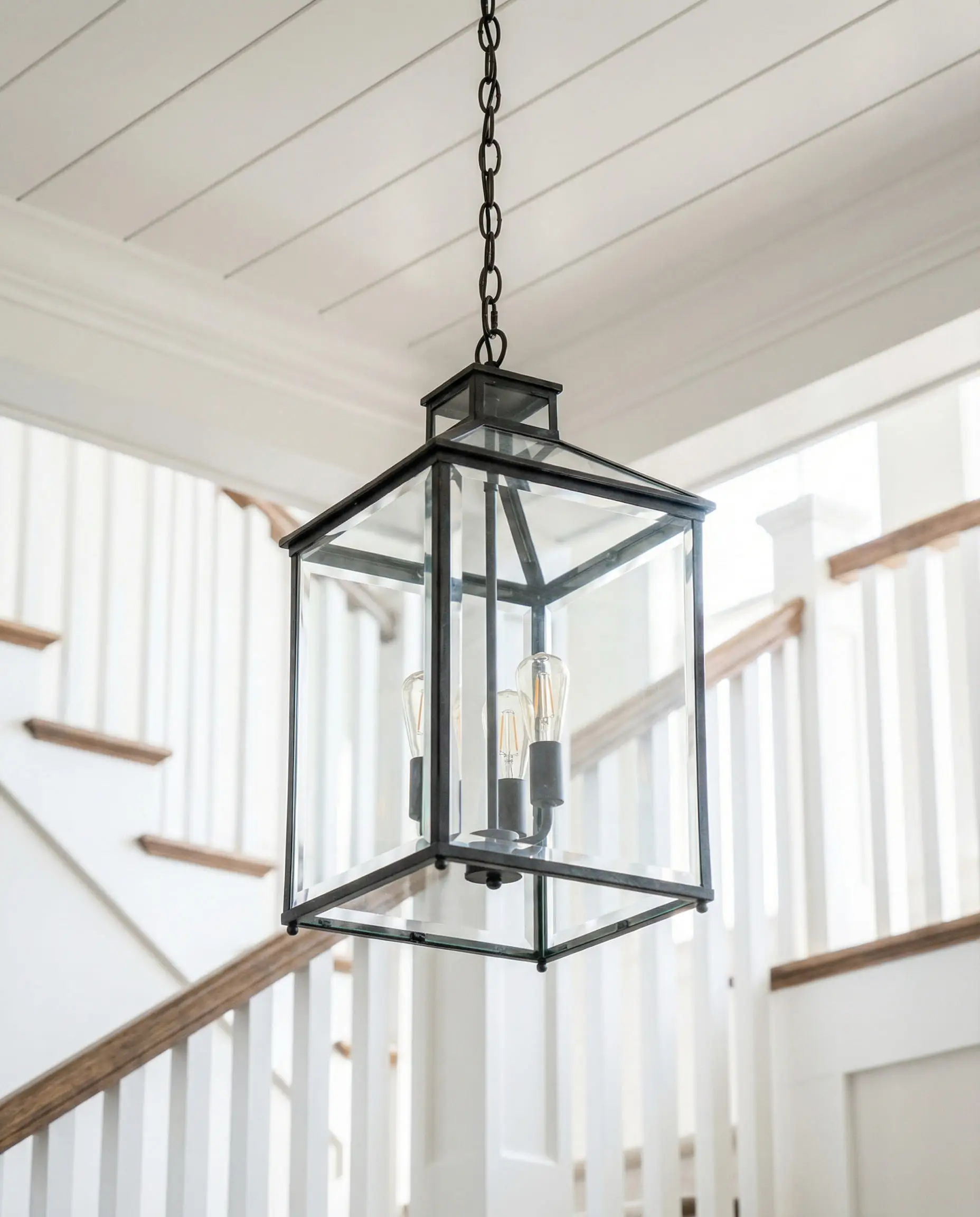 Iron and glass geometric lantern pendant light hanging in a bright craftsman entryway with a staircase in the background.