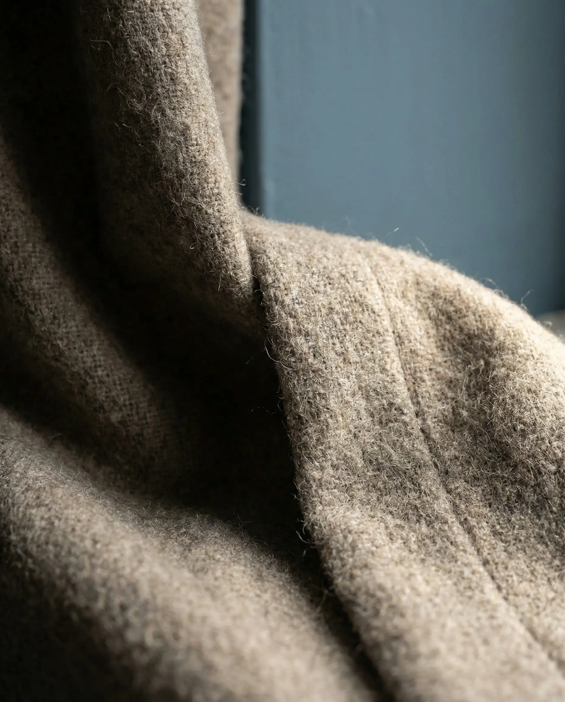 Close-up texture shot of heavy greige wool curtain fabric draped against a slate blue wall.