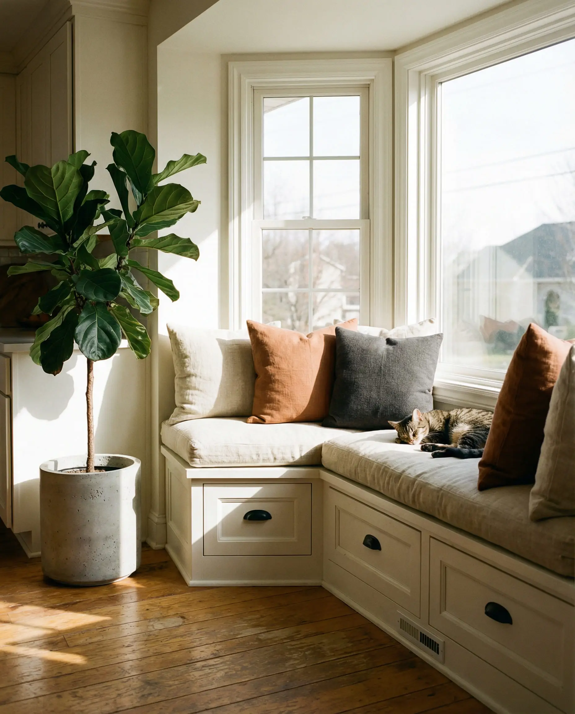 Sun-drenched kitchen window seat painted warm white with drawers featuring black pulls and linen pillows.