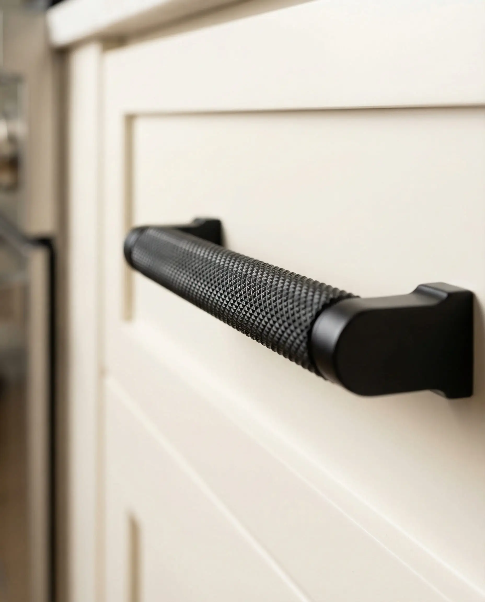 Macro photography detail of a diamond-knurled matte black cabinet pull mounted on a warm white flat-panel drawer front.