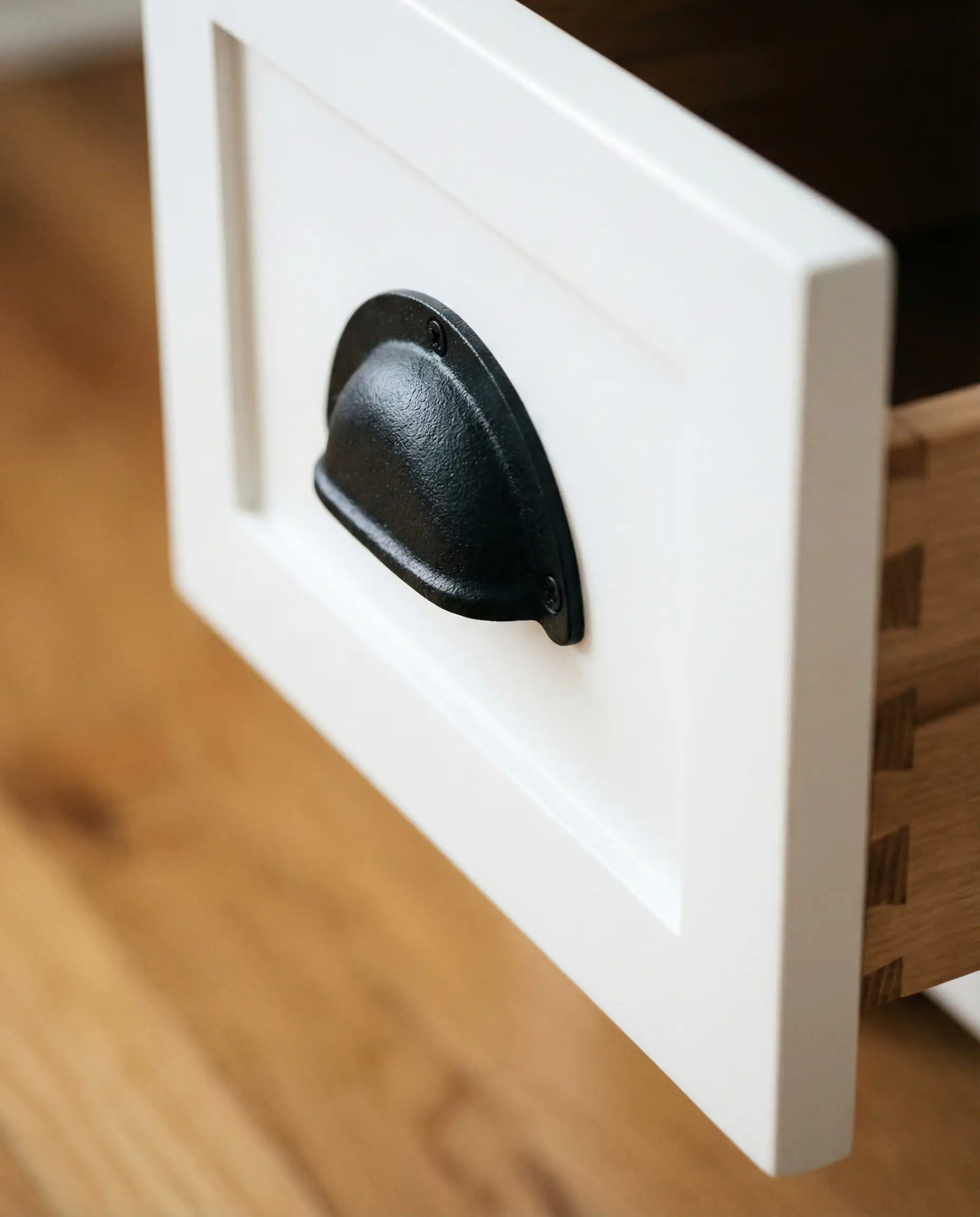 Close-up detail of a rustic matte black bin cup pull installed on a soft white shaker style drawer front.