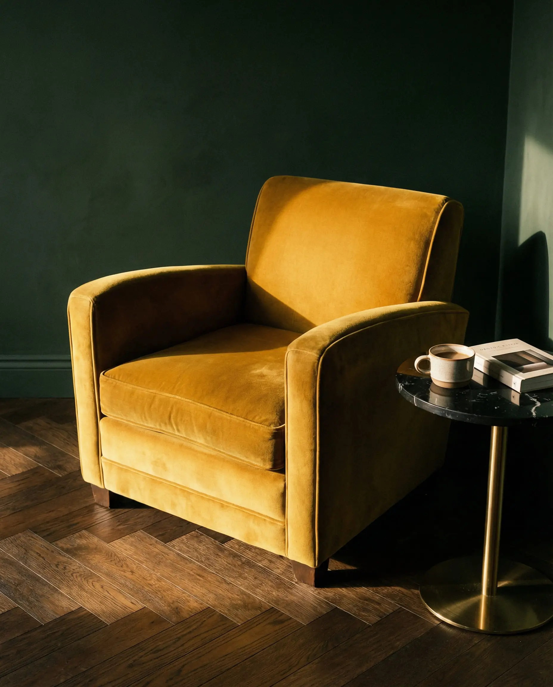 An interior vignette featuring a muddy mustard yellow velvet armchair on a herringbone wood floor bathed in warm golden light.