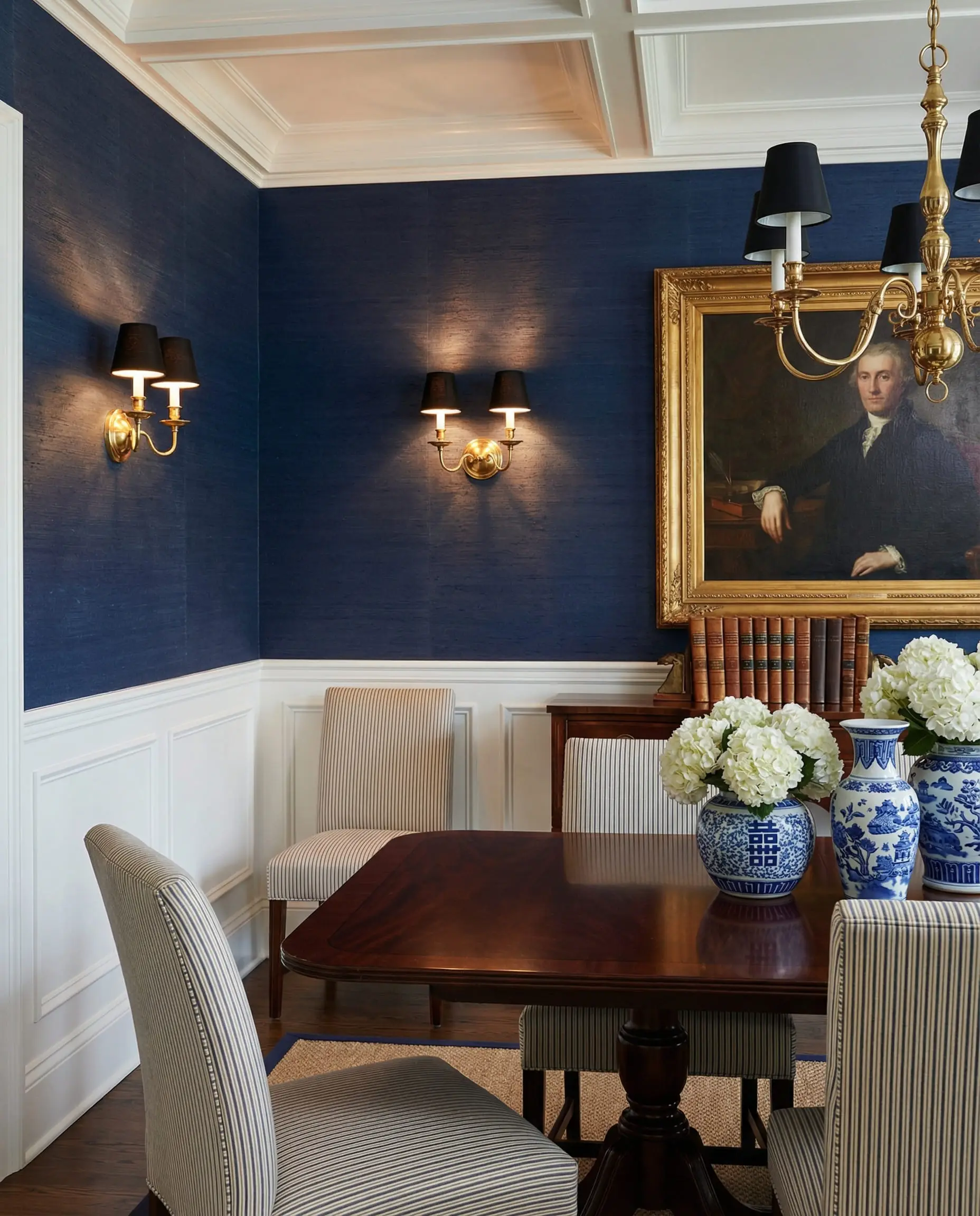 Sophisticated dining room with navy blue grasscloth wallpaper, white wainscoting, and brass wall sconces.