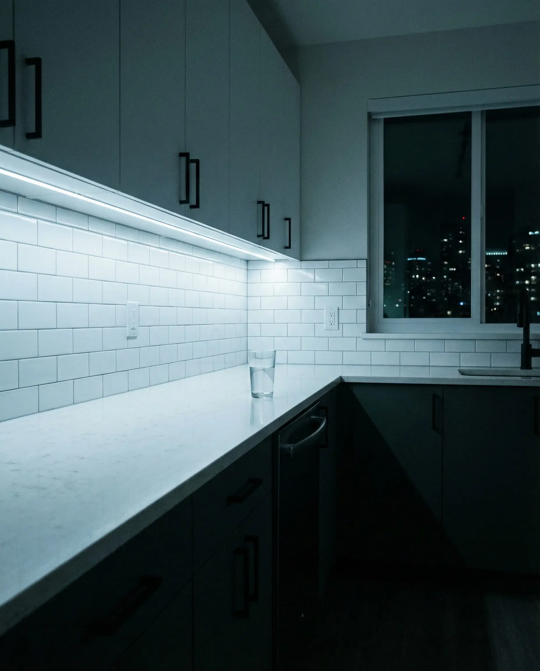 Minimalist kitchen at night with under-cabinet LED lighting illuminating white cabinets and black handles.