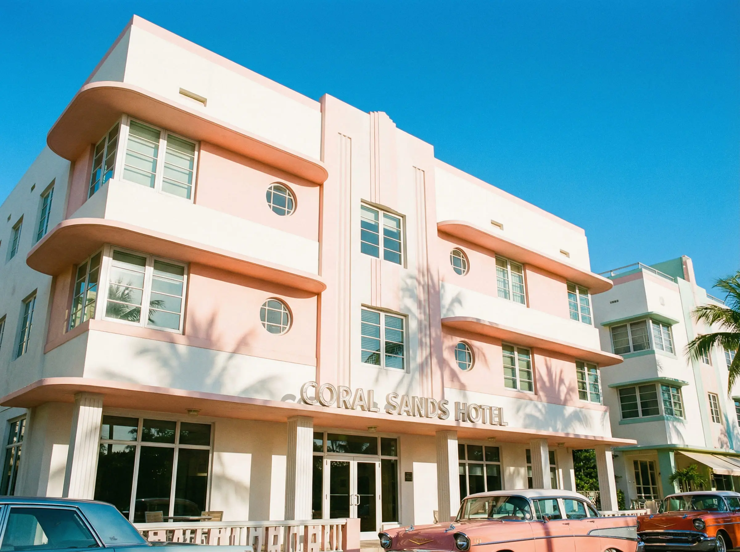 Exterior of a classic Miami Art Deco building featuring pastel pink walls, white relief details, and palm tree shadows against a blue sky.