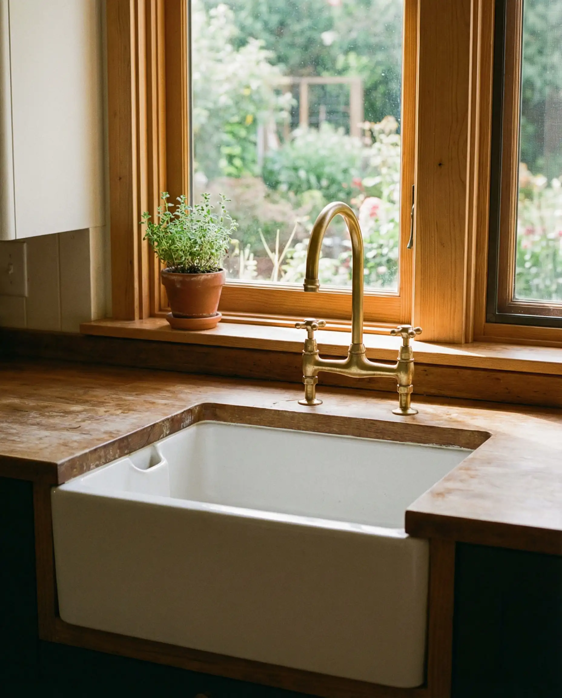 Fireclay farmhouse sink with an unlacquered brass bridge faucet in a white kitchen.