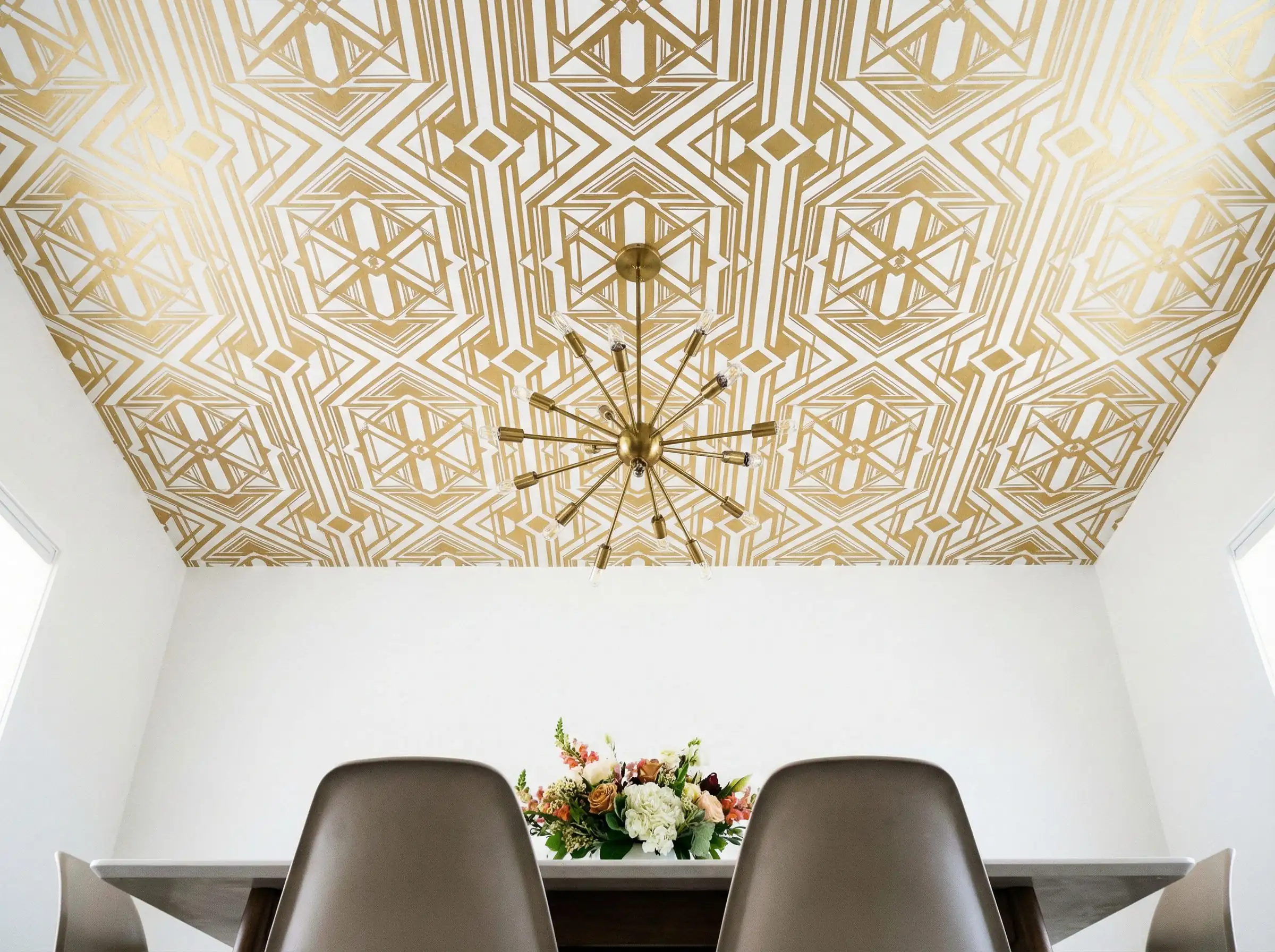 Low-angle shot of a dining room with a geometric gold and white wallpapered ceiling and a modern sputnik chandelier.