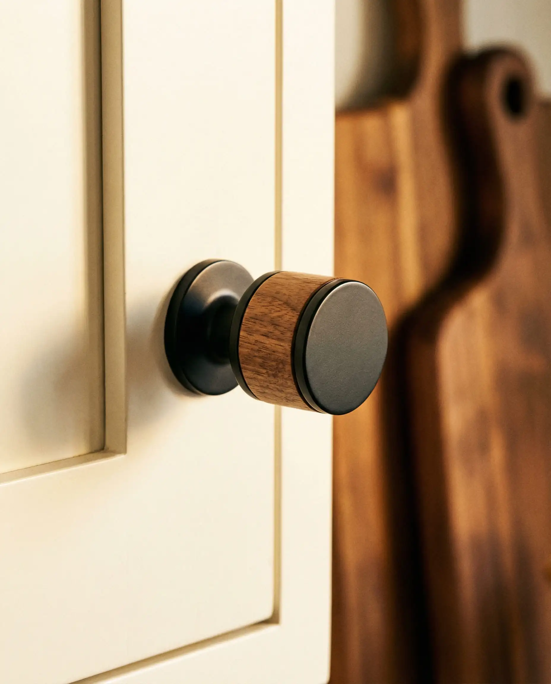 Detail of a cabinet knob made of natural walnut wood and matte black metal installed on a creamy white cabinet.