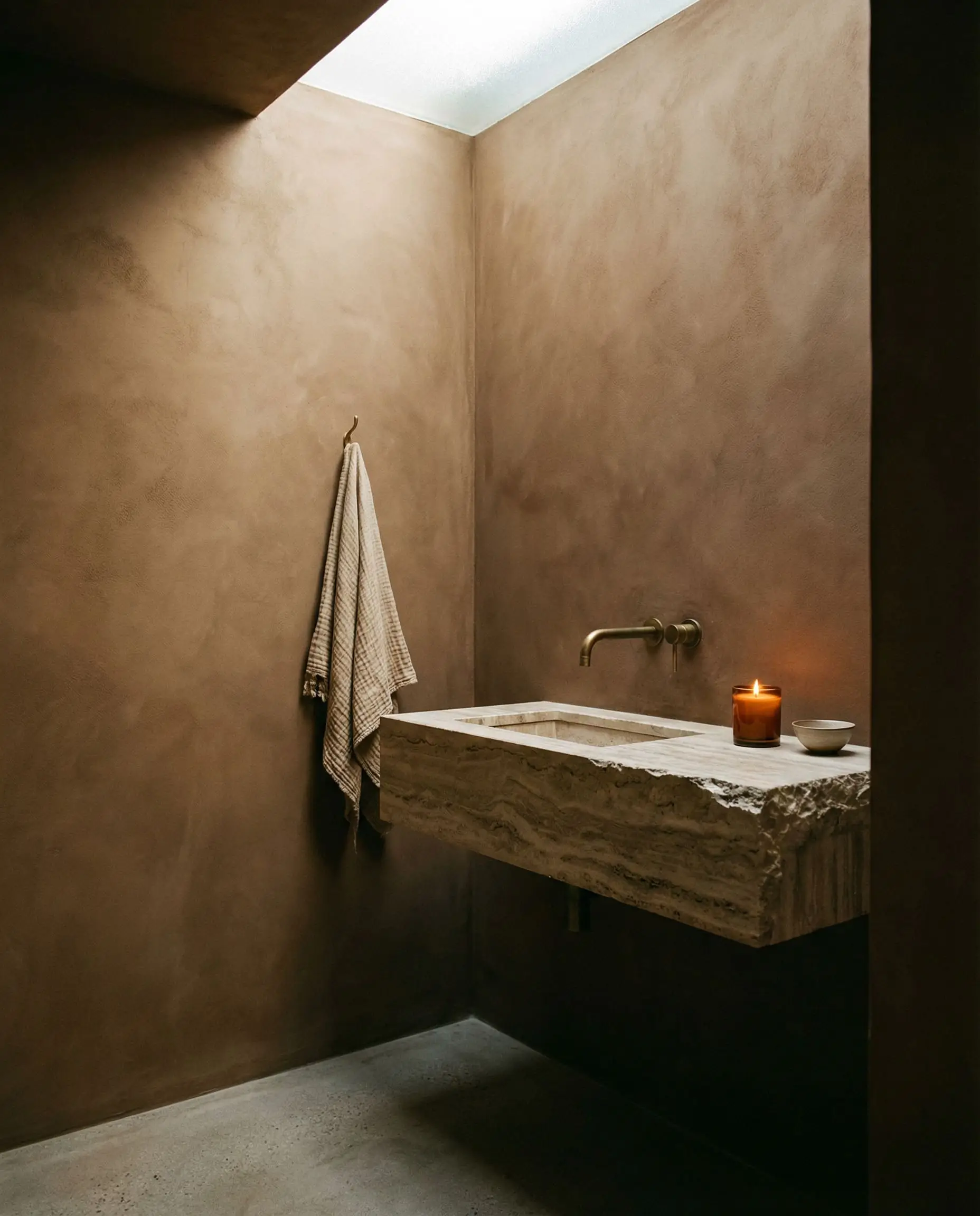 Warm brown textured wall paint creates an earthy wabi-sabi guest bathroom showcasing a floating travertine sink and unlacquered brass faucet.