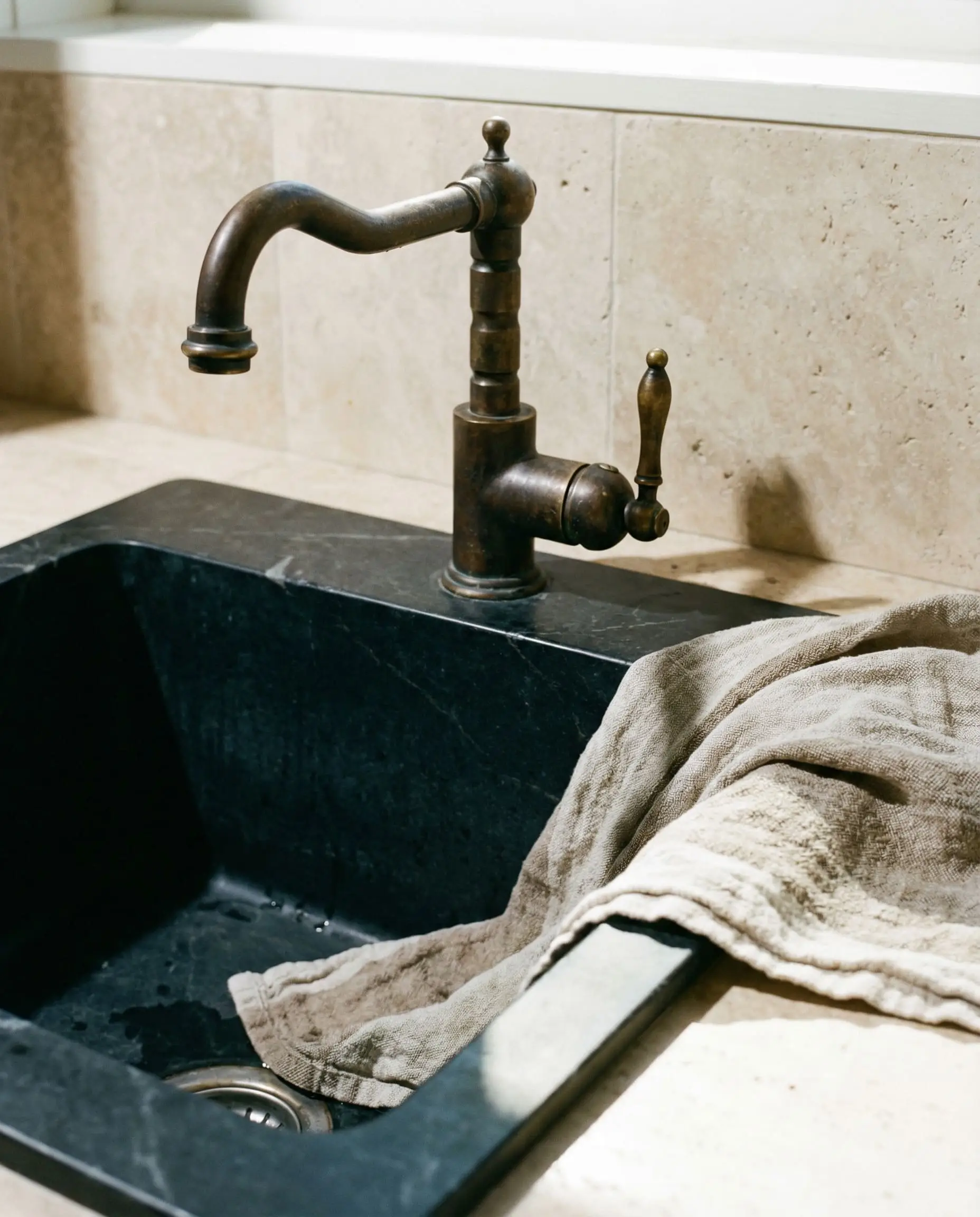 An unlacquered brass faucet with a dark aged patina sits above a deep matte soapstone sink basin against a honed travertine backsplash.