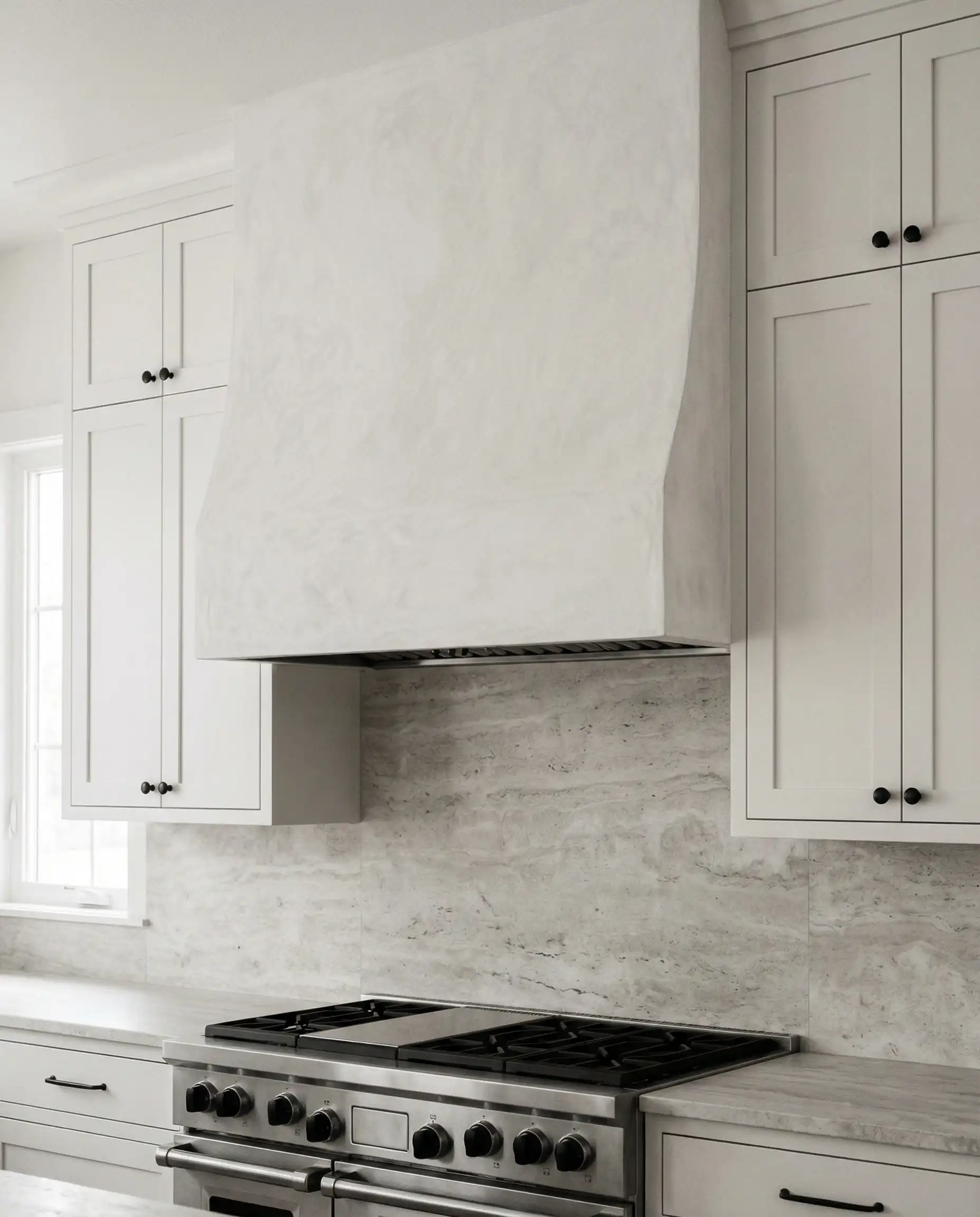 Vertical shot of a custom white Venetian plaster range hood flanked by white cabinets with simple black knobs.