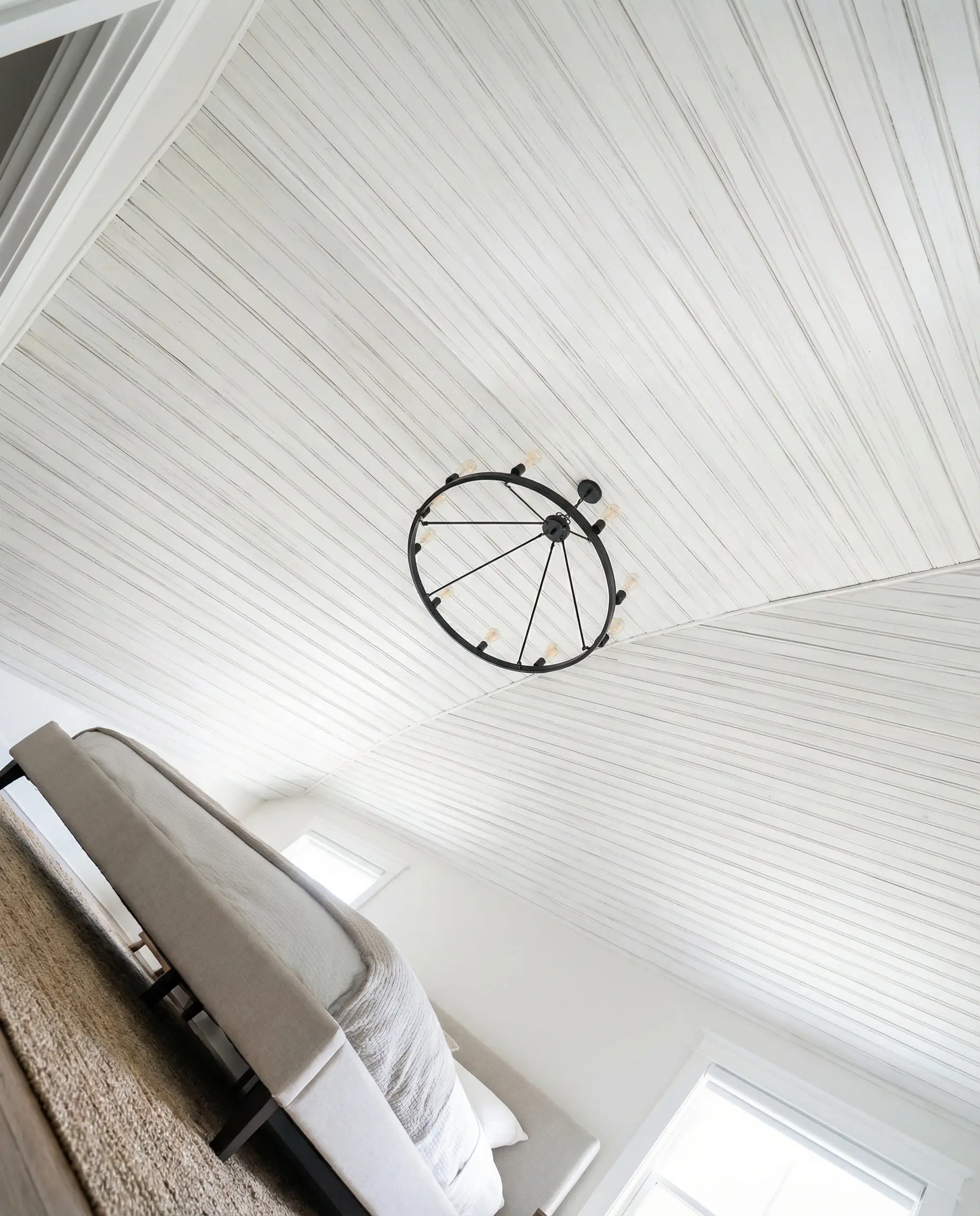 Bedroom ceiling clad in white tongue and groove paneling with an iron chandelier.