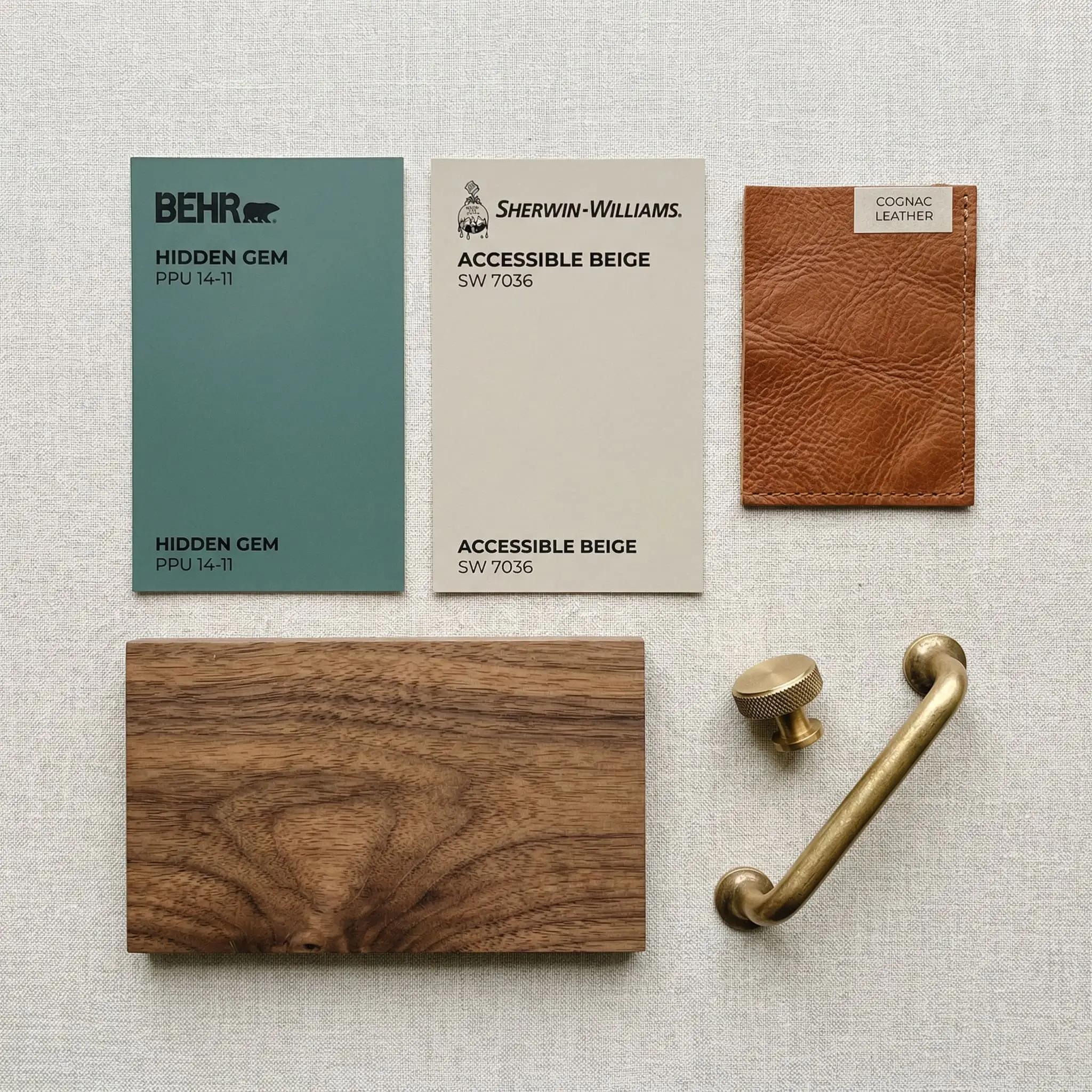 A painted color card of Behr Hidden Gem (Hex #596D69) rests alongside Sherwin-Williams Accessible Beige, a natural walnut wood sample, cognac leather, and brass hardware.