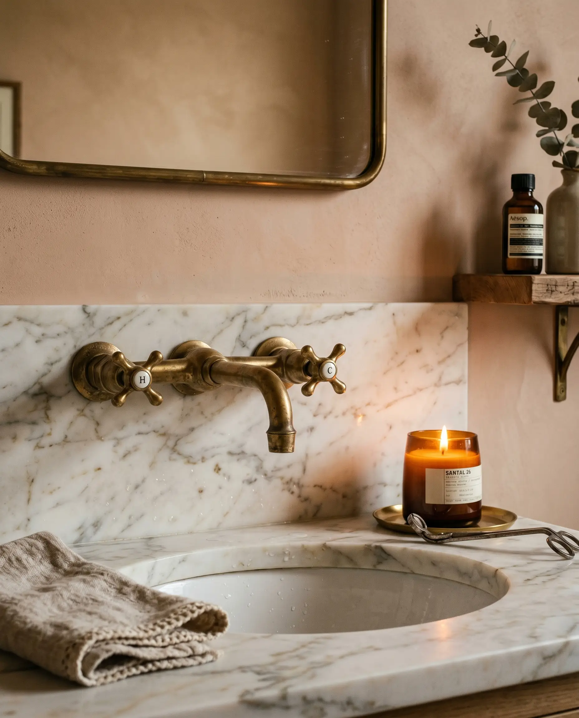 An unlacquered brass faucet sits over a honed Calacatta Gold marble sink alongside a luxury linen hand towel and a lit amber glass candle.