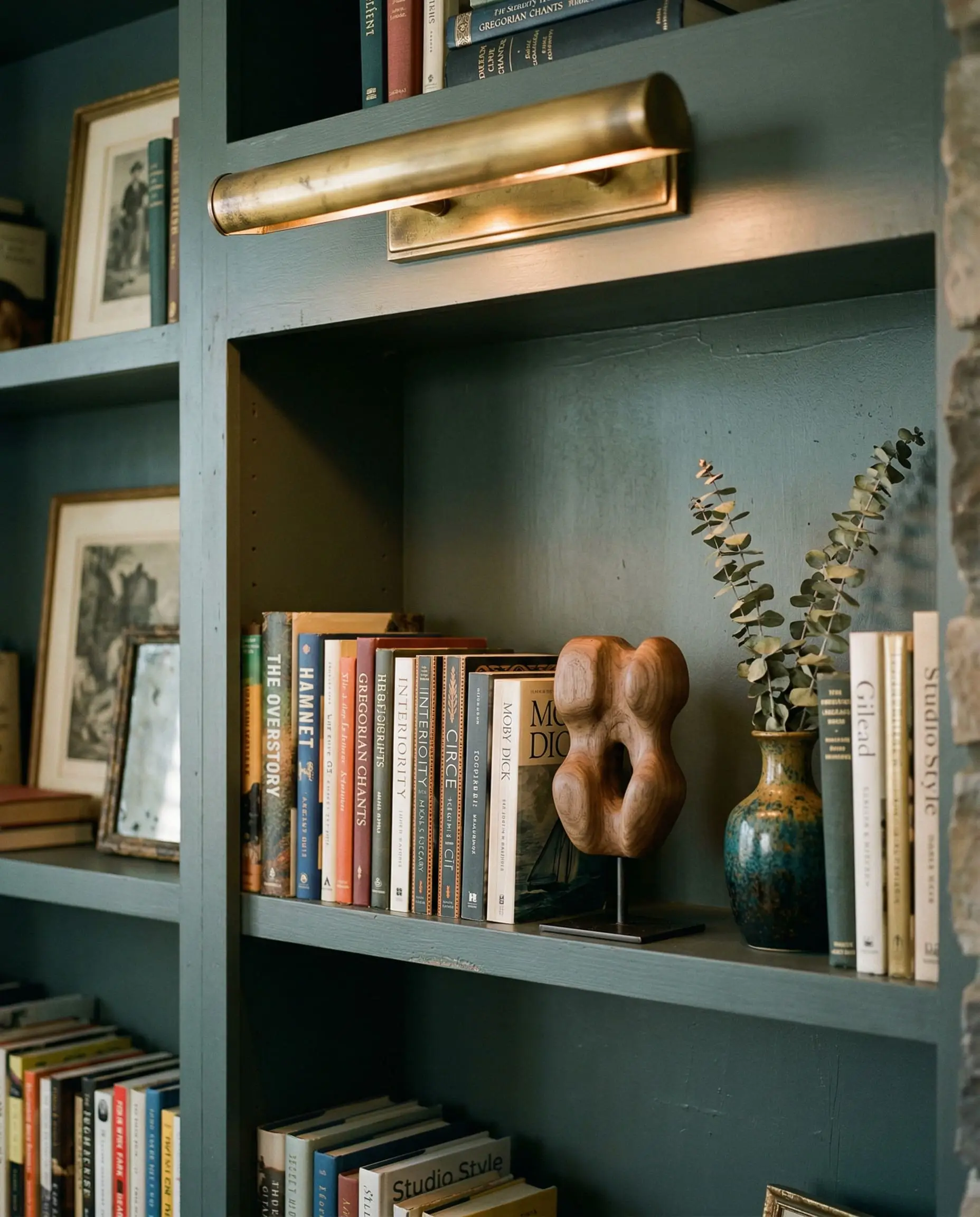 An unlacquered brass picture light casts a warm, localized glow over colorful book spines and a natural walnut sculpture on dark green built-in shelving.