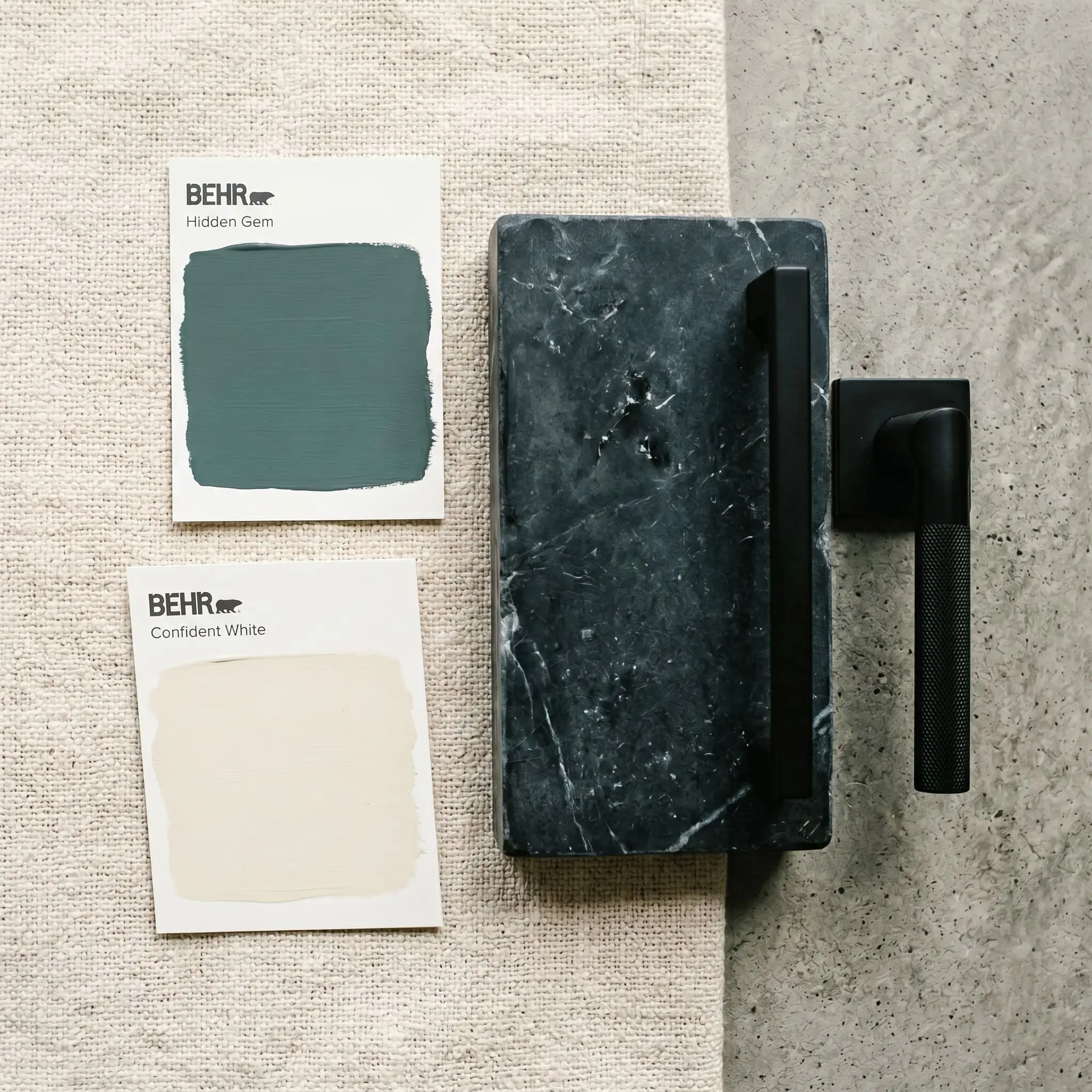 A smoky jade paint swatch sits beside Behr Confident White, heavy honed soapstone, matte black metal fixtures, and natural linen fabric.