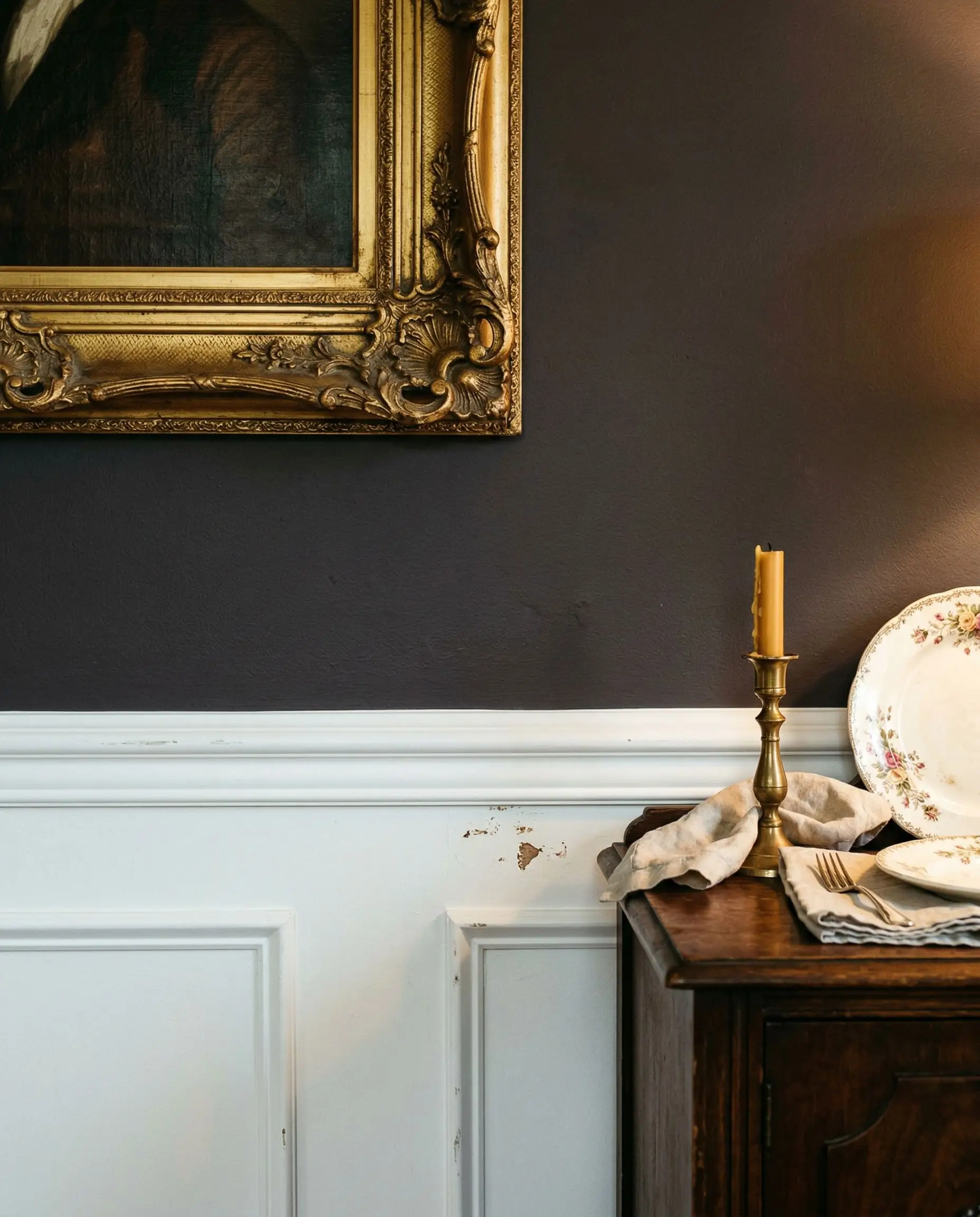 The edge of an ornate gilded picture frame hangs on a dark plum-charcoal upper wall that sharply meets crisp white wainscoting under warm ambient light.
