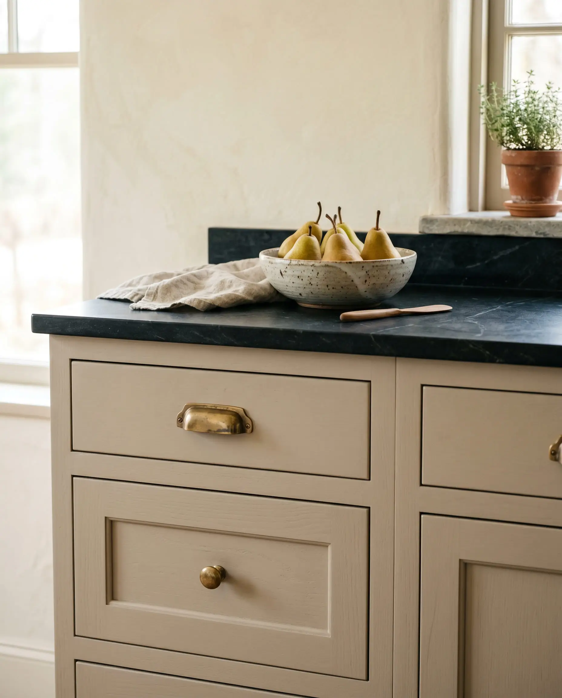 Lower kitchen drawers painted in an earthy mushroom feature unlacquered brass cabinet hardware beneath a matte-black honed soapstone countertop holding a ceramic bowl of pears.