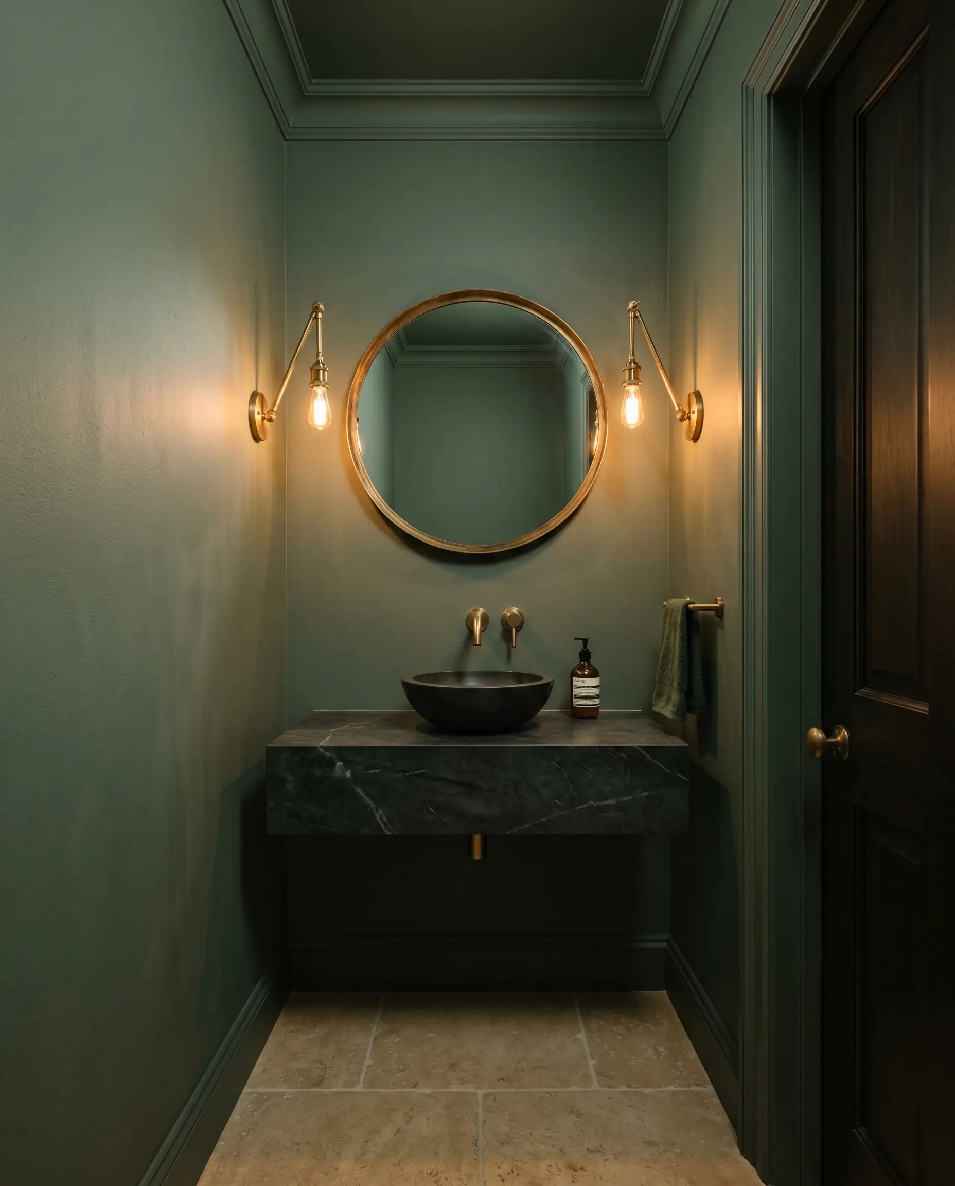 A windowless powder room color-drenched in Farrow & Ball Green Smoke (Hex #6f7b71) features a dark soapstone floating vanity and warm unlacquered brass wall sconces.