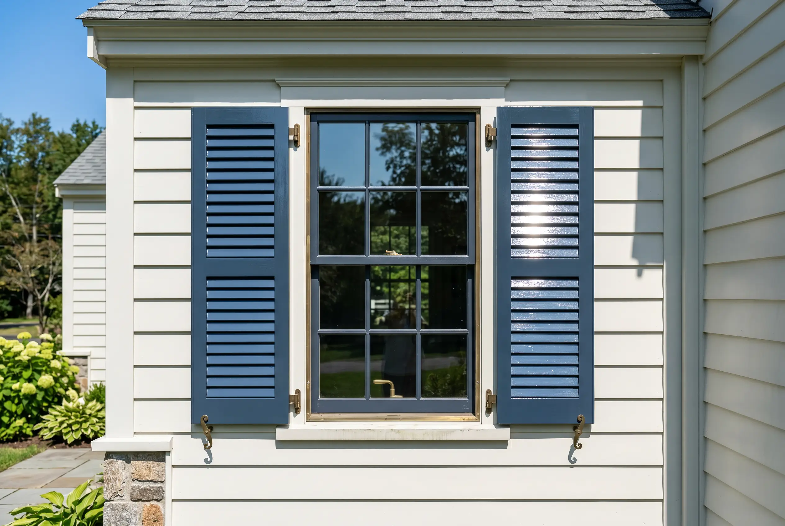 High-gloss exterior window shutters reflect bright sunlight against creamy white house siding and unlacquered brass exterior hardware.
