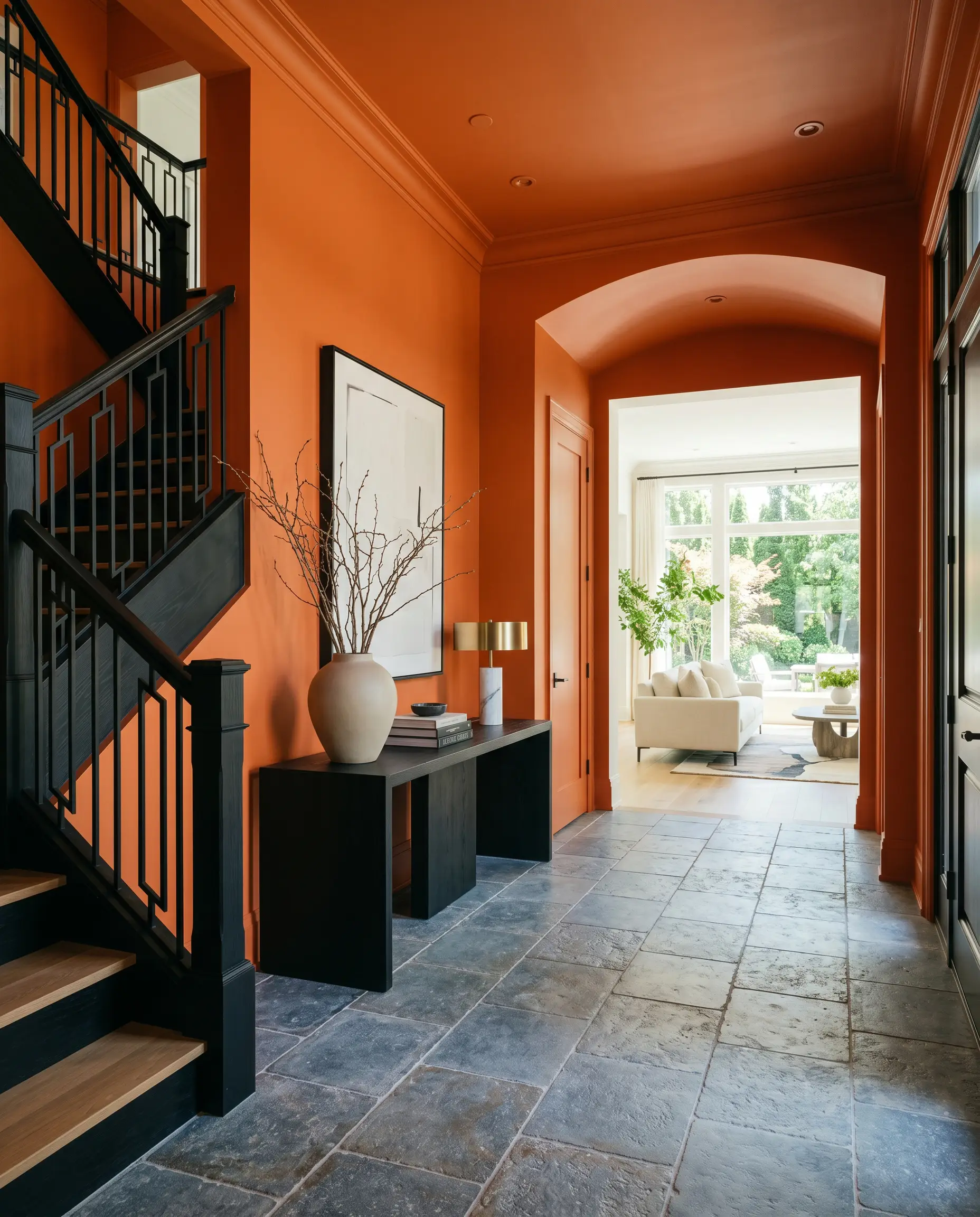 An ebonized wood console table sits on tumbled Belgian bluestone flooring in a vivid warm orange entryway that opens into a crisp white living room.